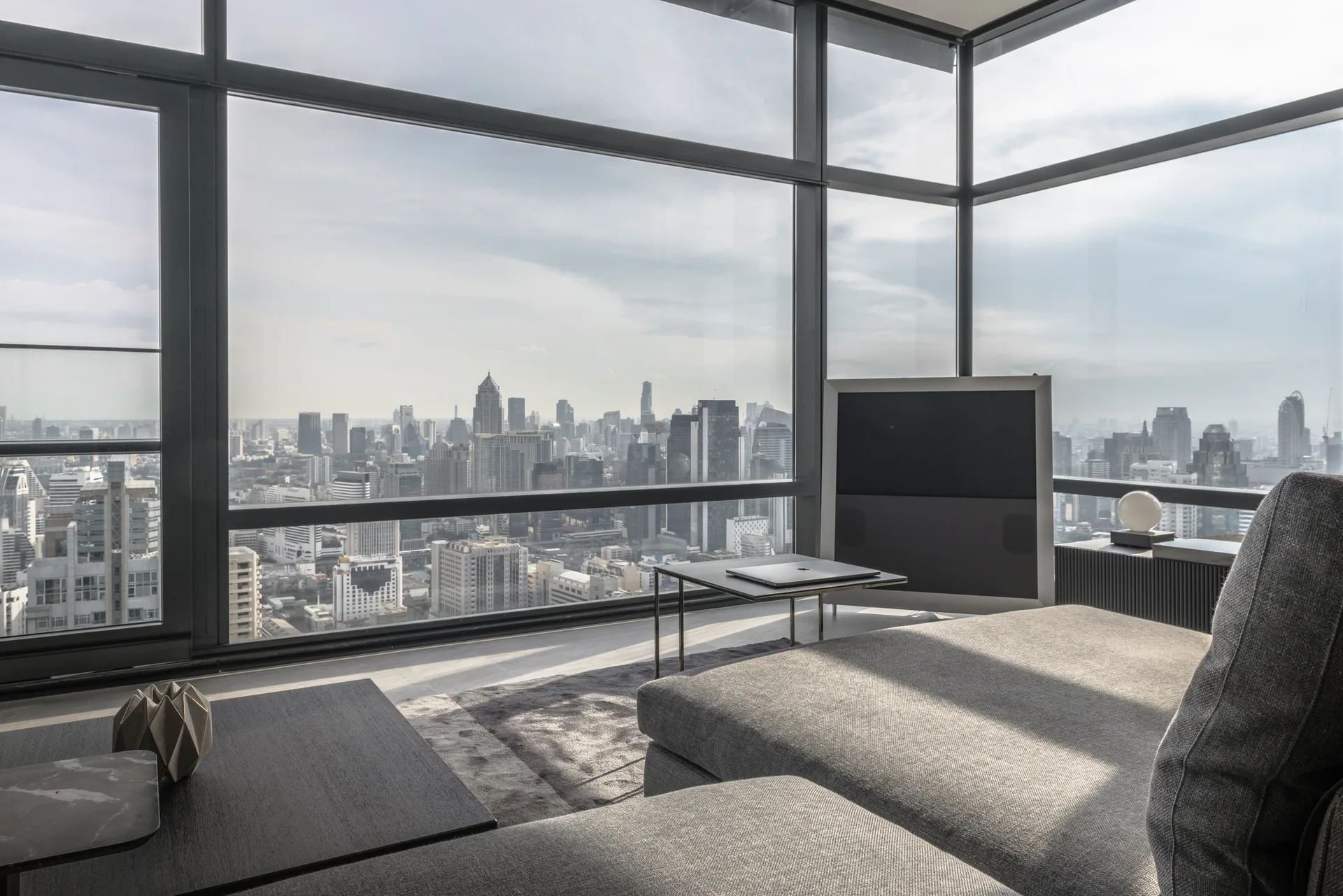 A minimalist living room features a grey sectional sofa, low metal tables, and large floor-to-ceiling windows overlooking a city skyline.