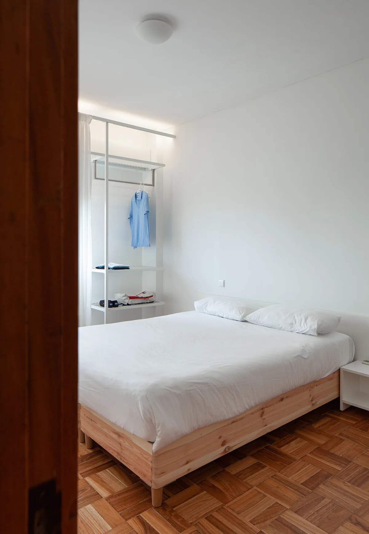 A minimalist bedroom features a light wood bed with white linens, a white open closet system, and a warm-toned parquet wood floor.