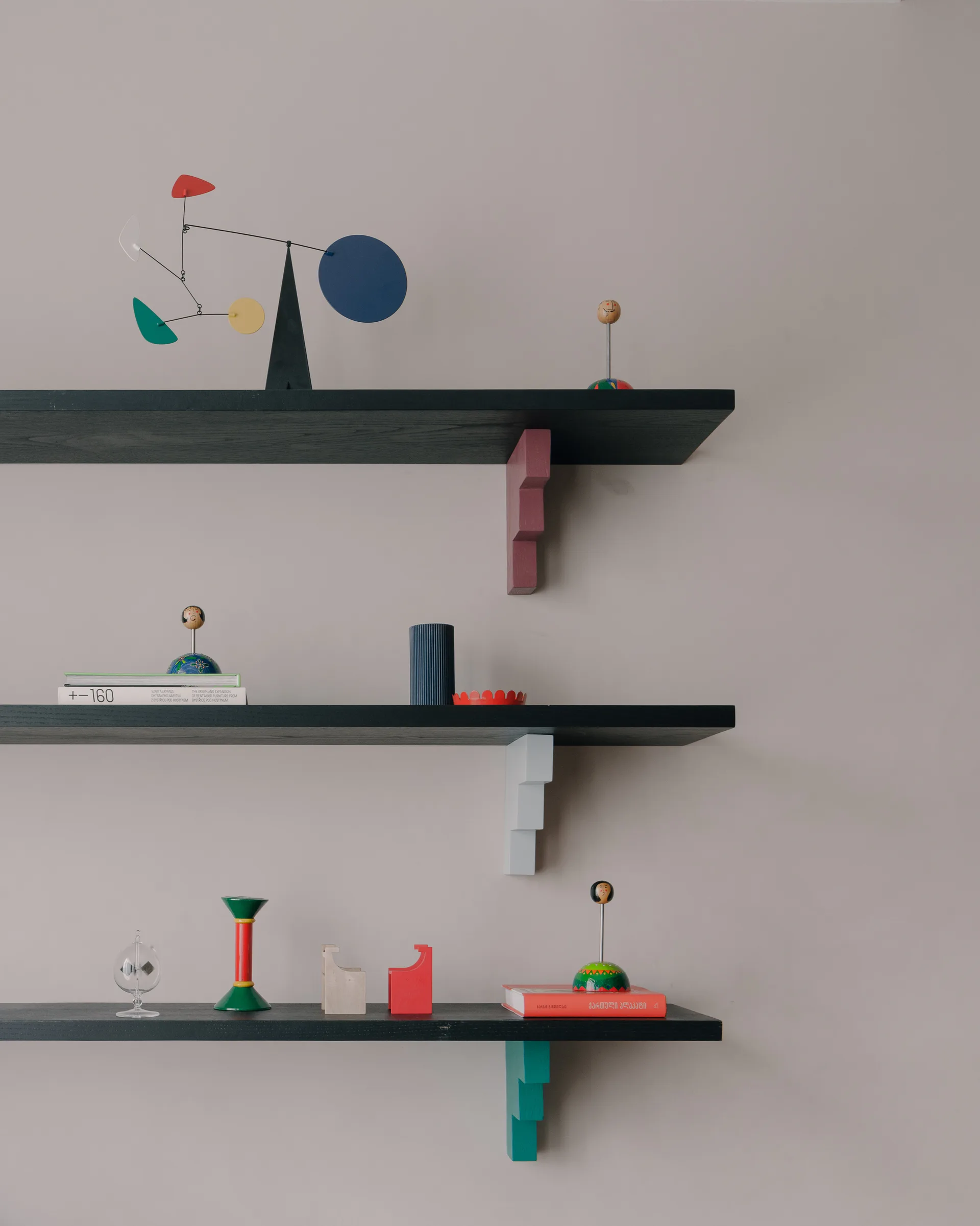 Three black wall-mounted shelves feature geometric brackets in pink, light blue, and teal, decorated with colorful sculptures, books, and figurines.