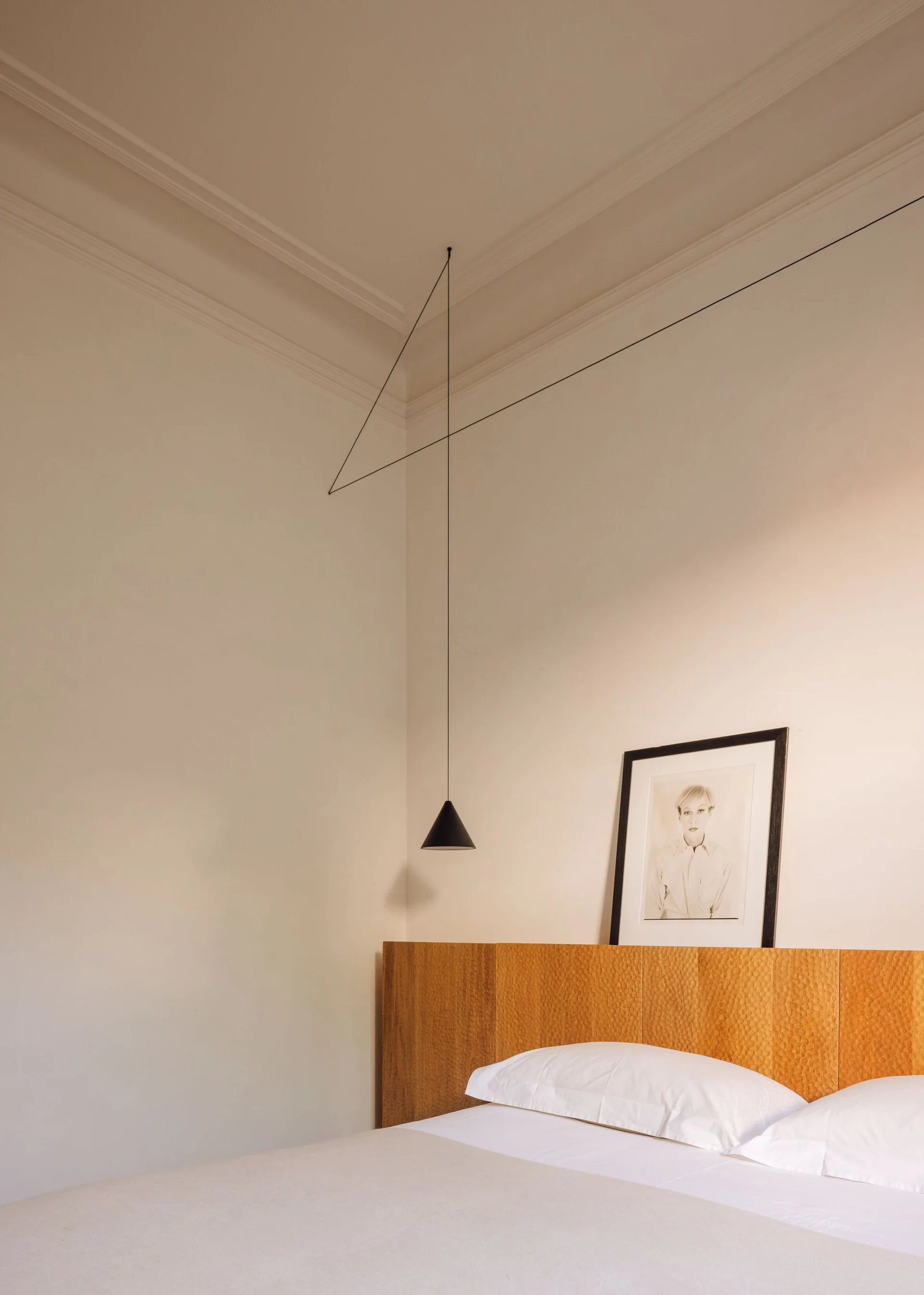 A minimalist bedroom featuring a textured wooden headboard, a white bed, a framed portrait, and a black wire pendant light.