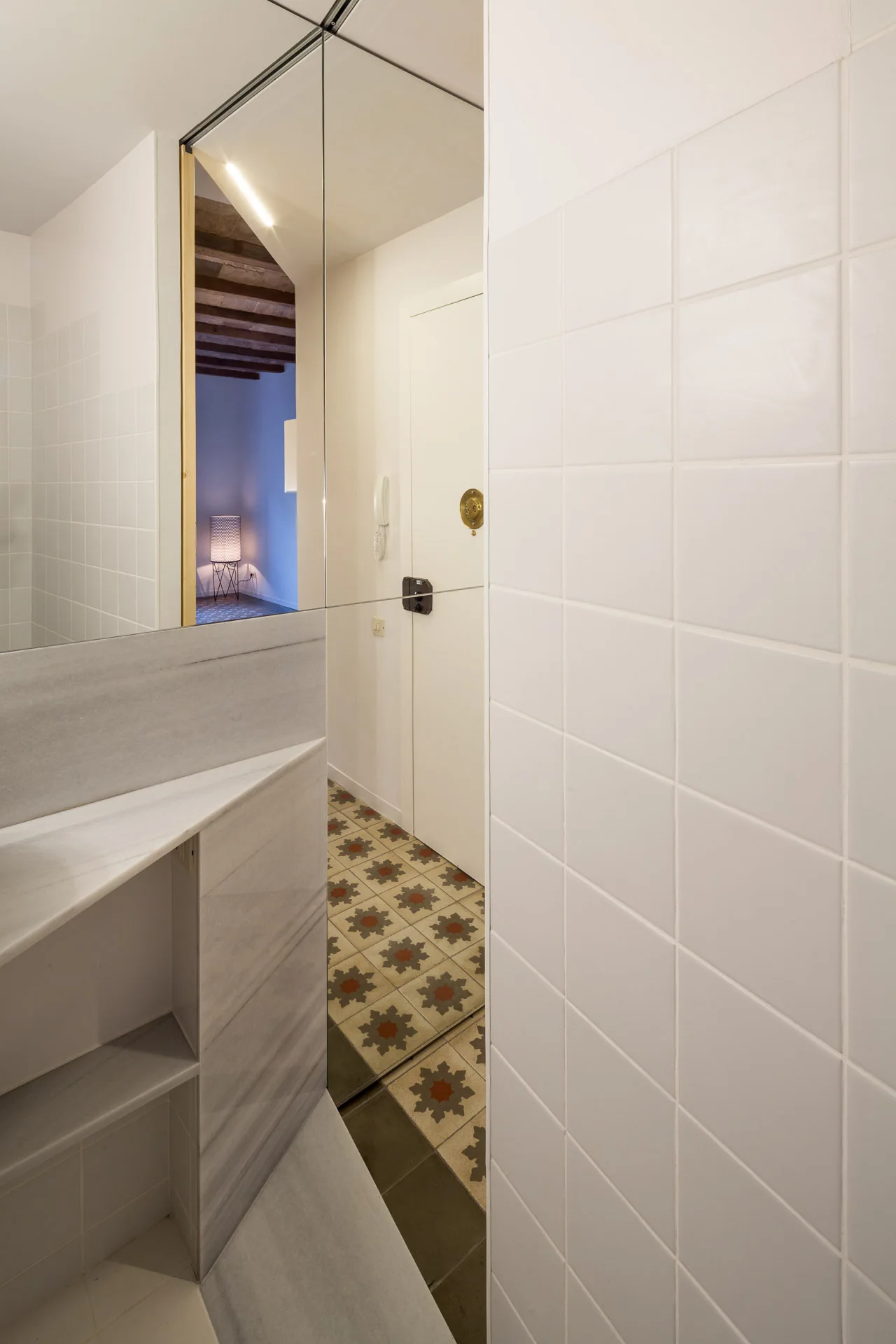 A modern bathroom features white tiled walls, a marble vanity shelf, and a tall mirror reflecting a hallway with geometric patterned floor tiles.