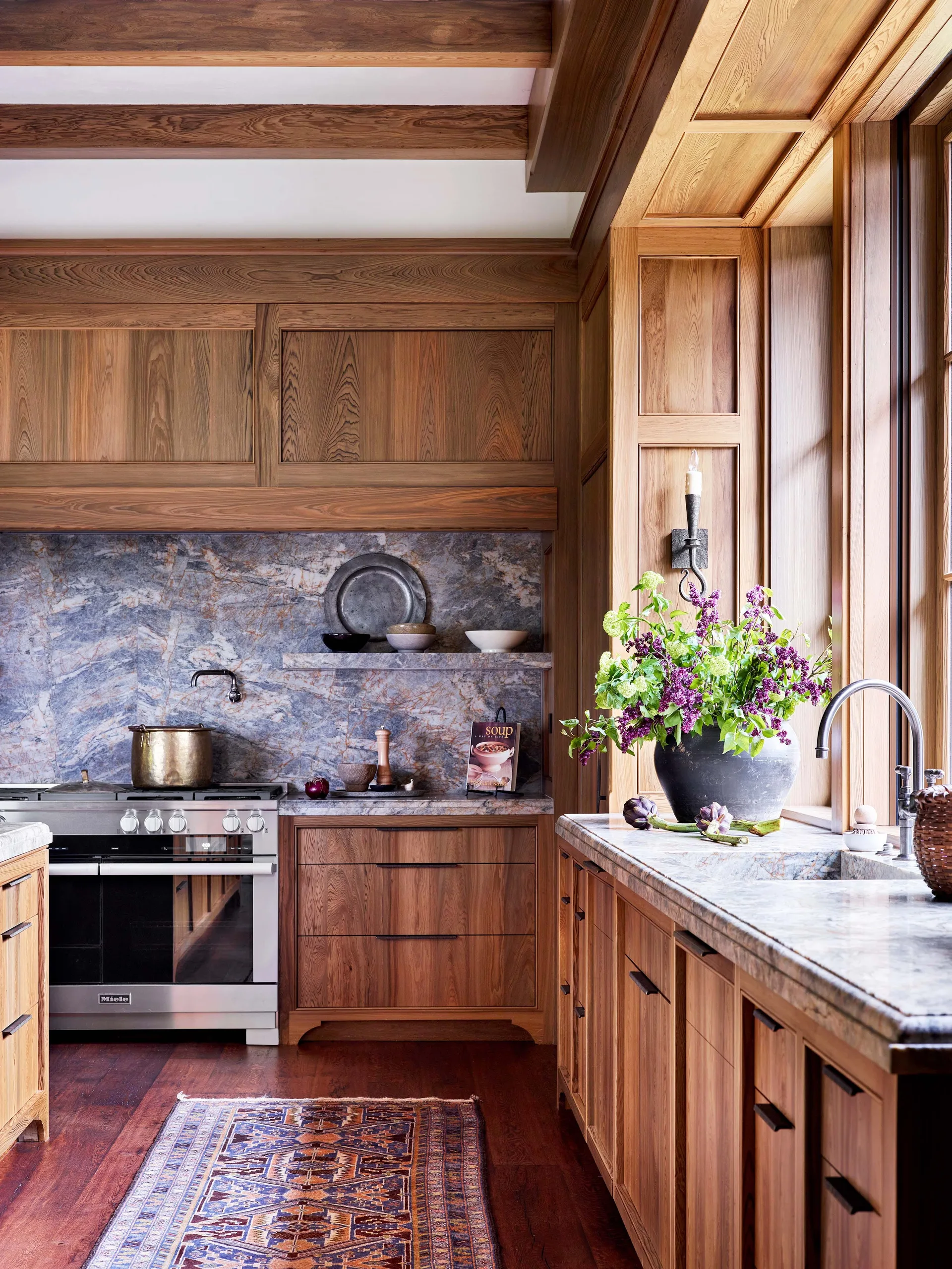 A kitchen features warm wood cabinetry, a heavily veined blue-grey stone backsplash, a stainless steel range, and a patterned runner rug on dark floors.