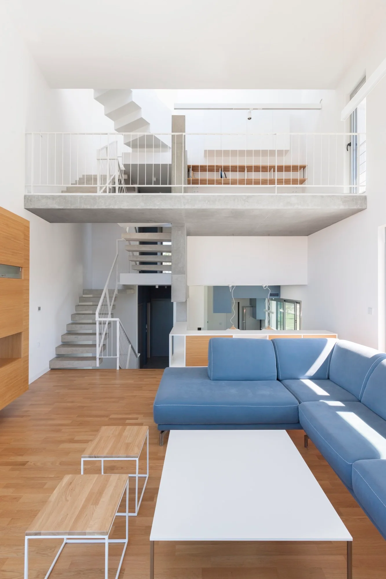 A modern loft living room features a blue sectional sofa, a white coffee table, and an architectural concrete staircase leading to a mezzanine.