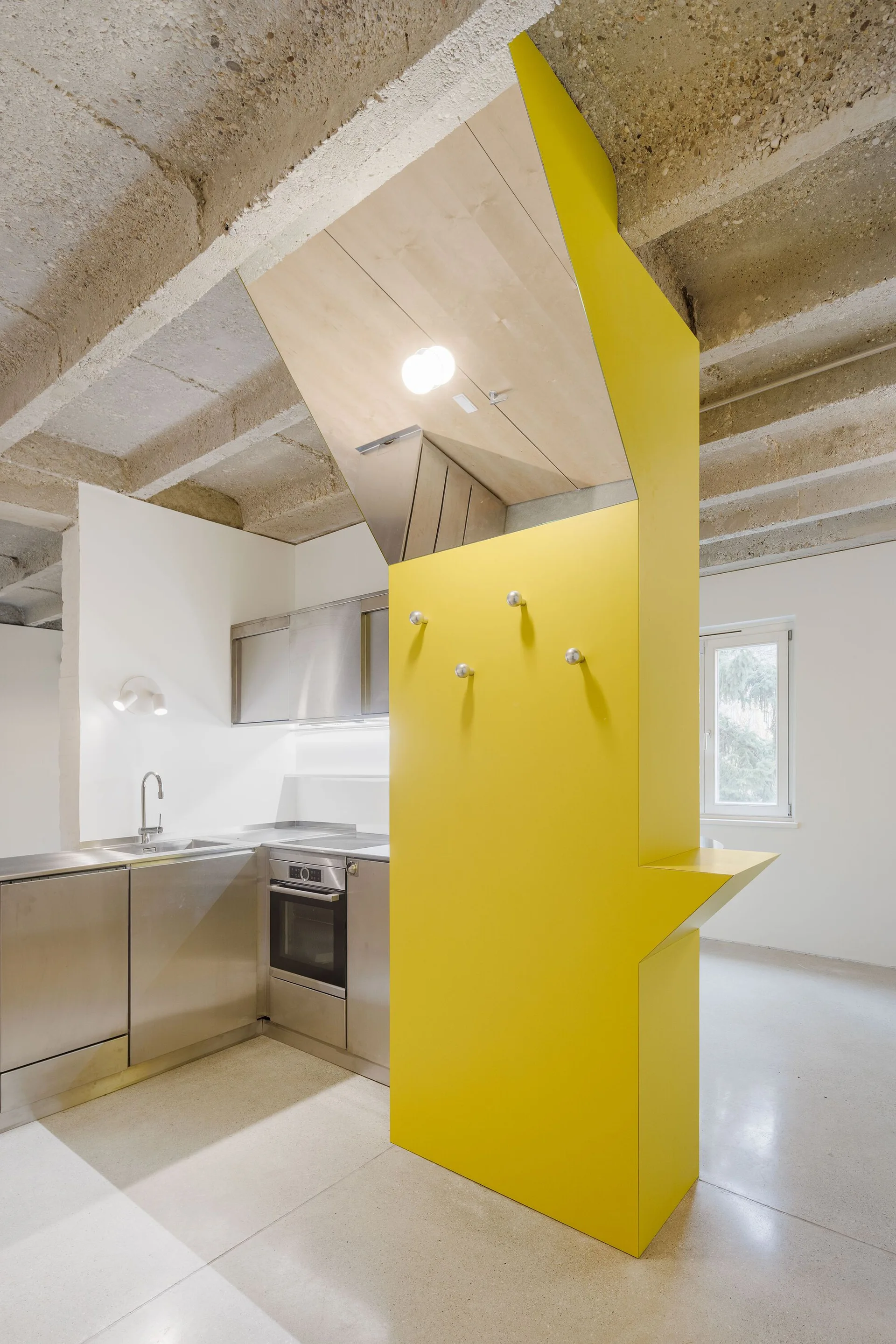 A modern kitchen features stainless steel cabinetry and a built-in oven beneath an exposed concrete ceiling, separated by a bright yellow room divider.