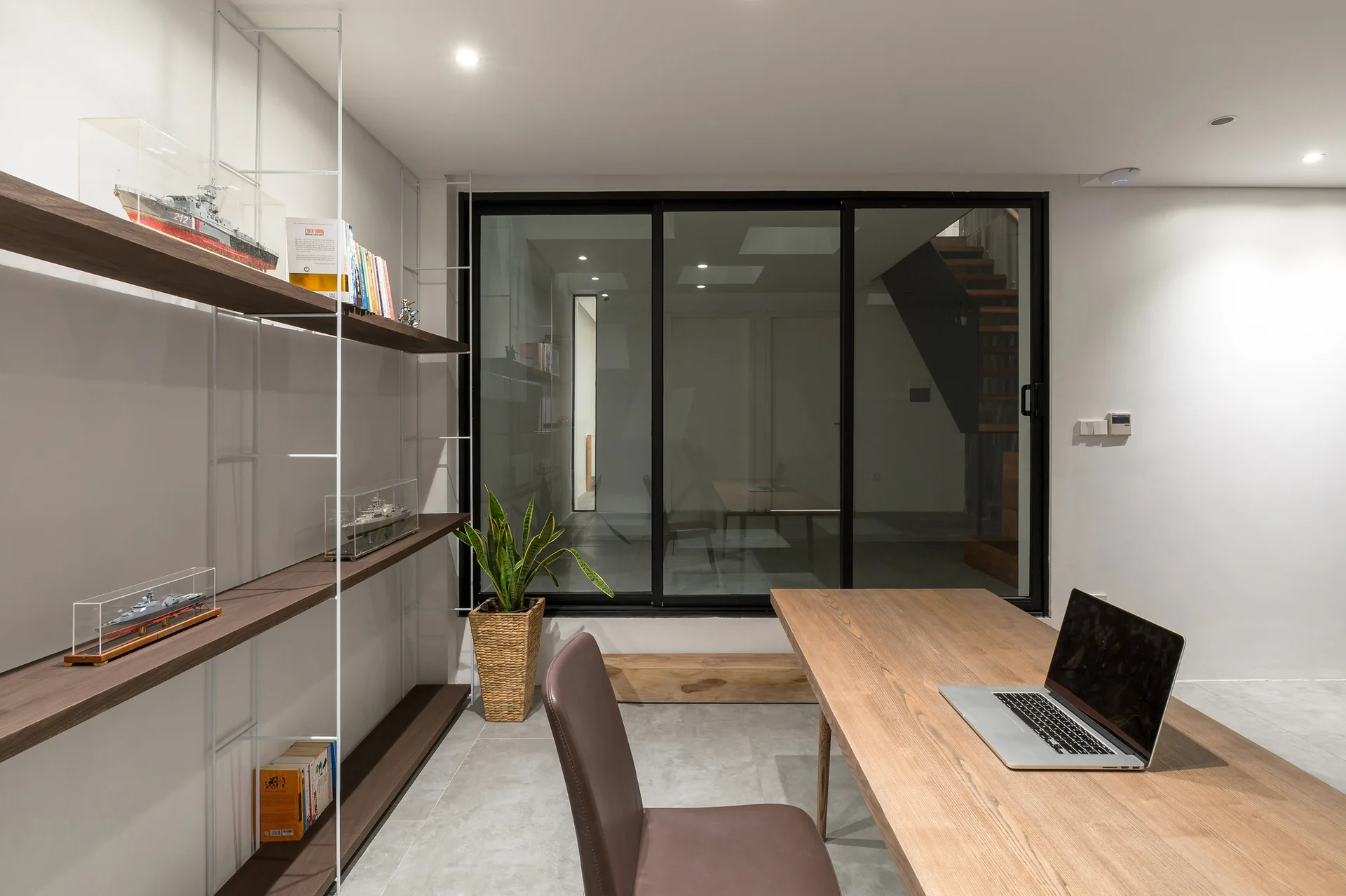 A modern home office with a minimalist wooden desk, a brown leather-like chair, and a white metal shelving unit beside a sliding glass partition.