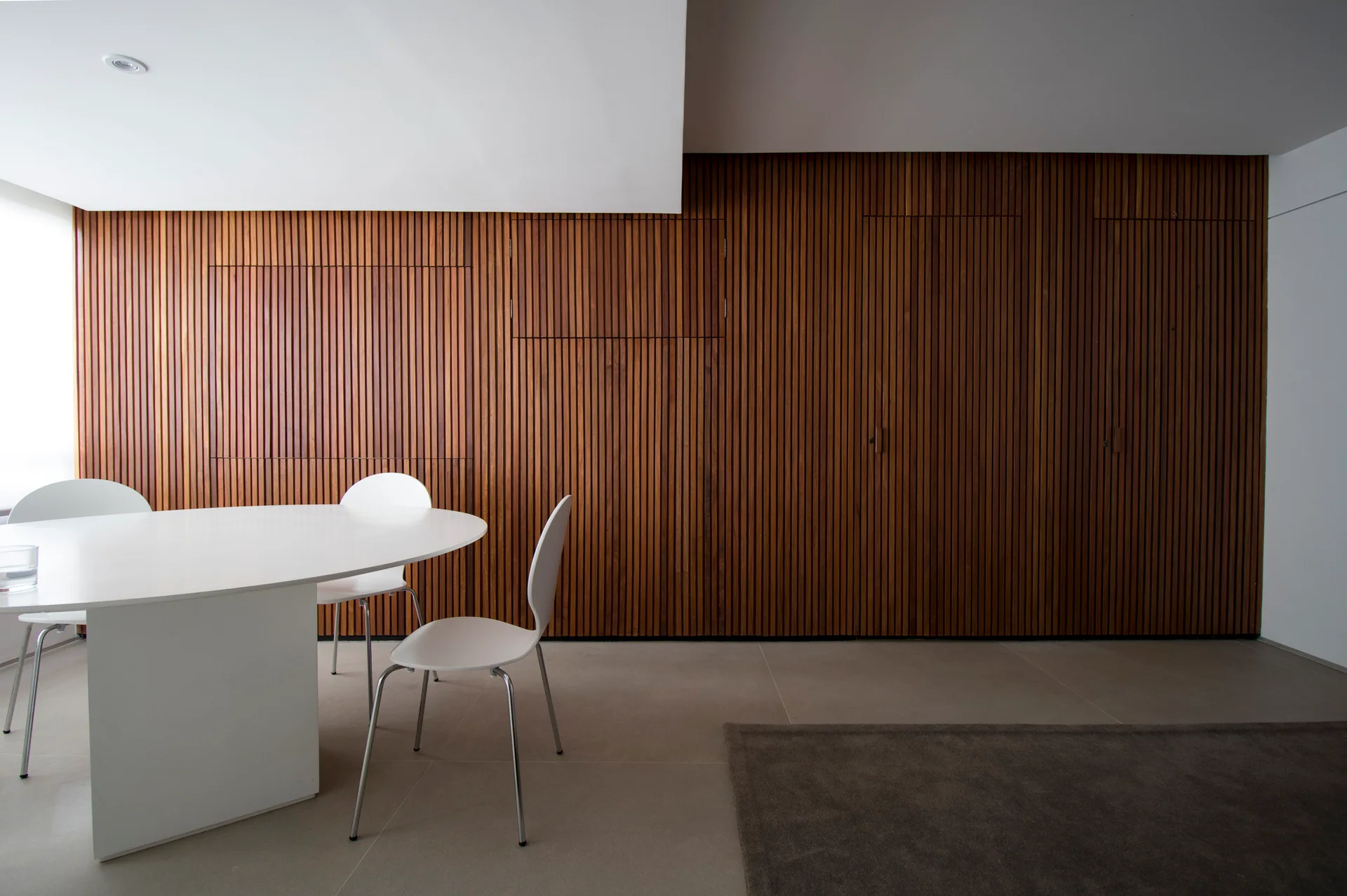A minimalist white oval dining table and matching chairs stand before a full wall of vertical wooden slats on a tiled floor.