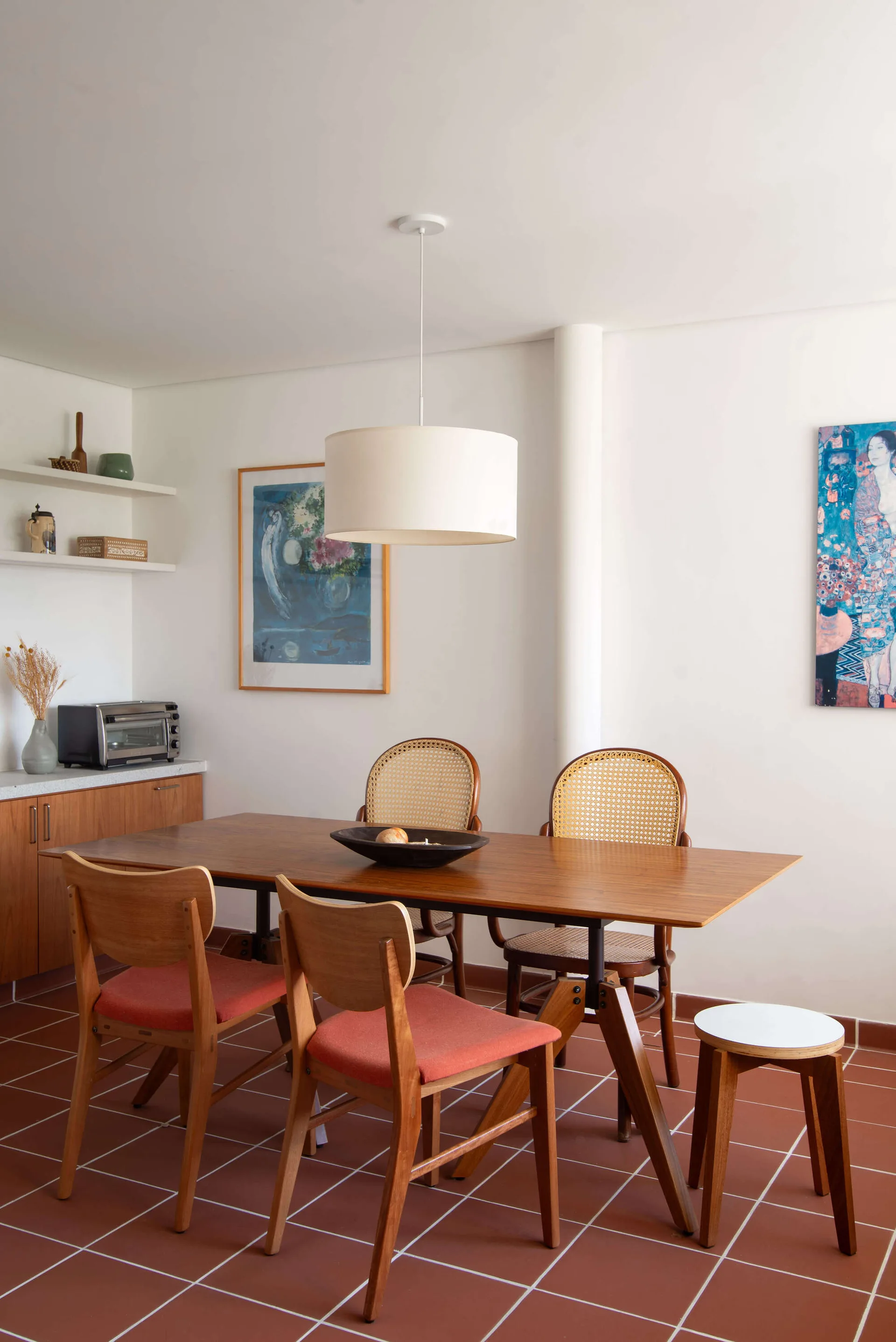 A wooden dining table and mixed chairs sit on terracotta tile flooring beneath a large white drum pendant light.