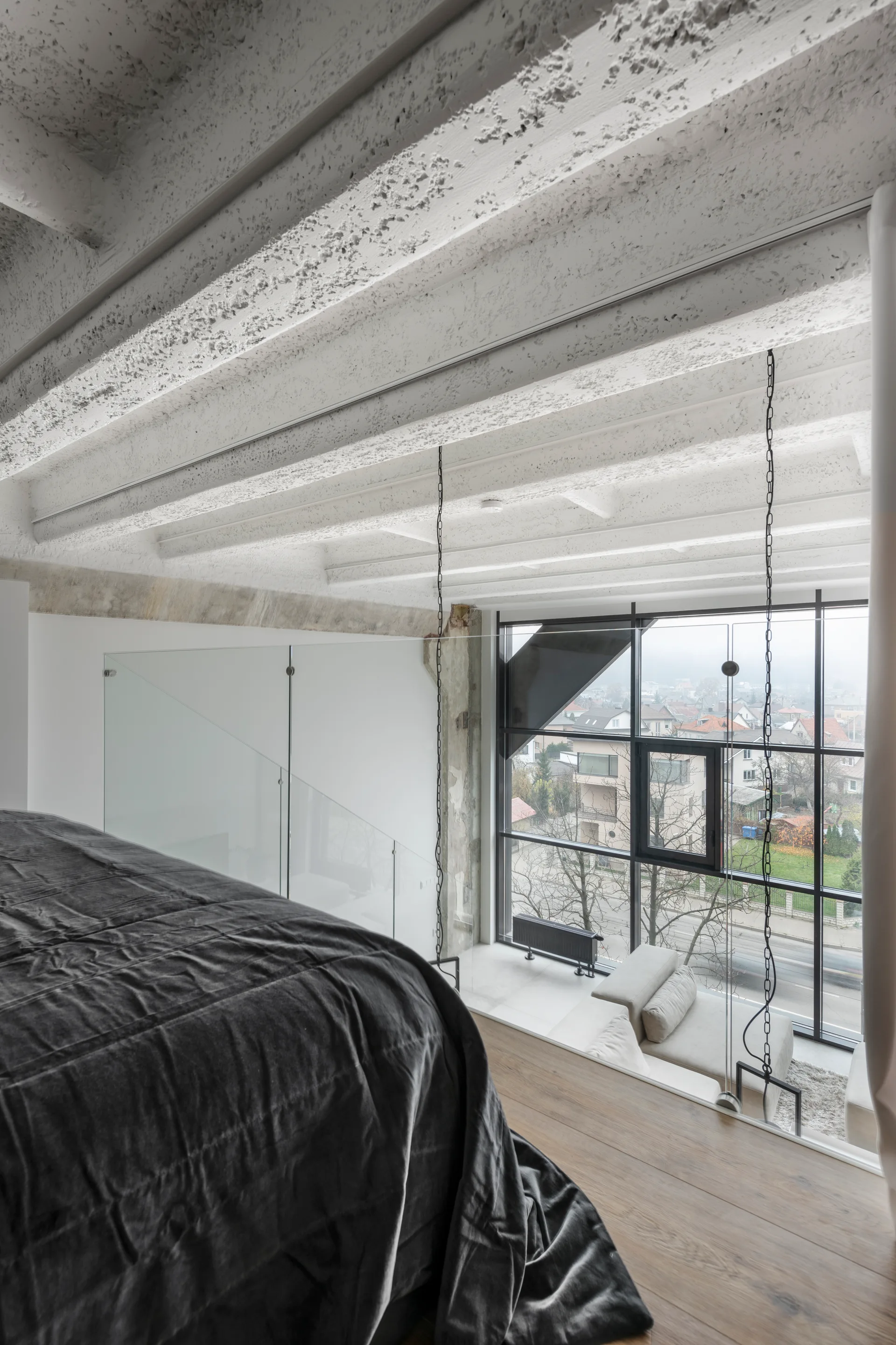 A loft bedroom mezzanine features a grey bed on wood flooring, textured concrete ceiling beams overhead, and a glass railing overlooking a tall window.