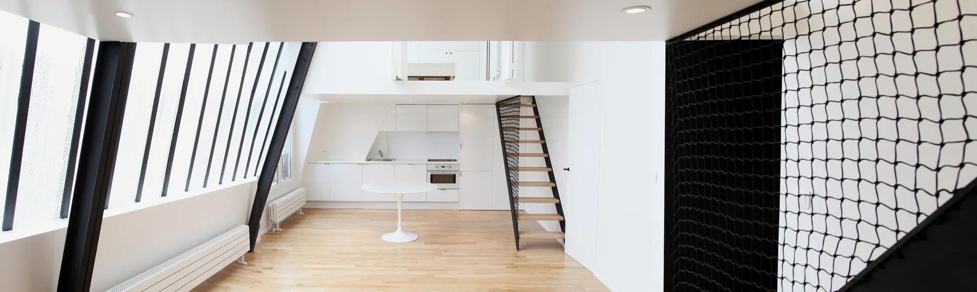 A modern loft interior featuring a white kitchen, a wooden staircase with black safety netting, a round pedestal table, and slanted windows.