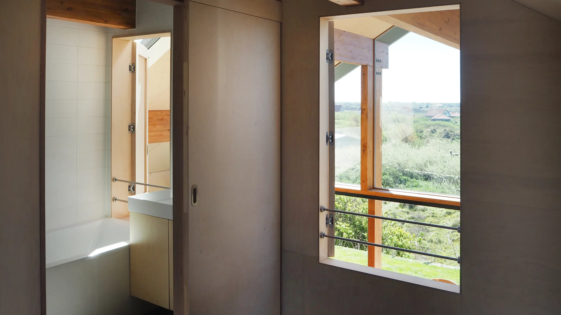 A minimalist cabin interior featuring light plywood walls, a pocket sliding door, a white bathtub, and a large window looking out at a grassy landscape.