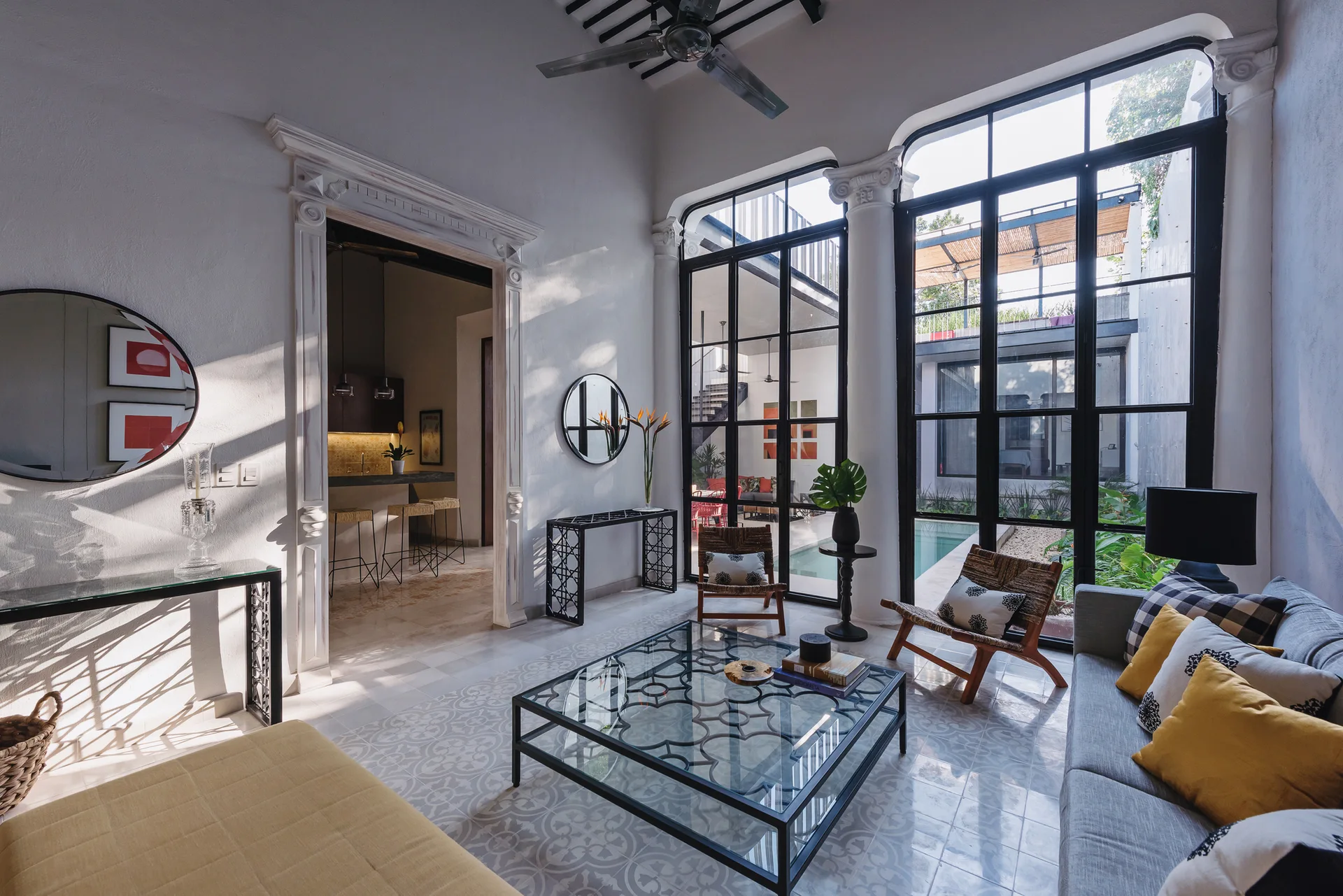A bright living room features large black steel-framed windows looking out to a pool courtyard, patterned floor tiles, and a grey sofa.