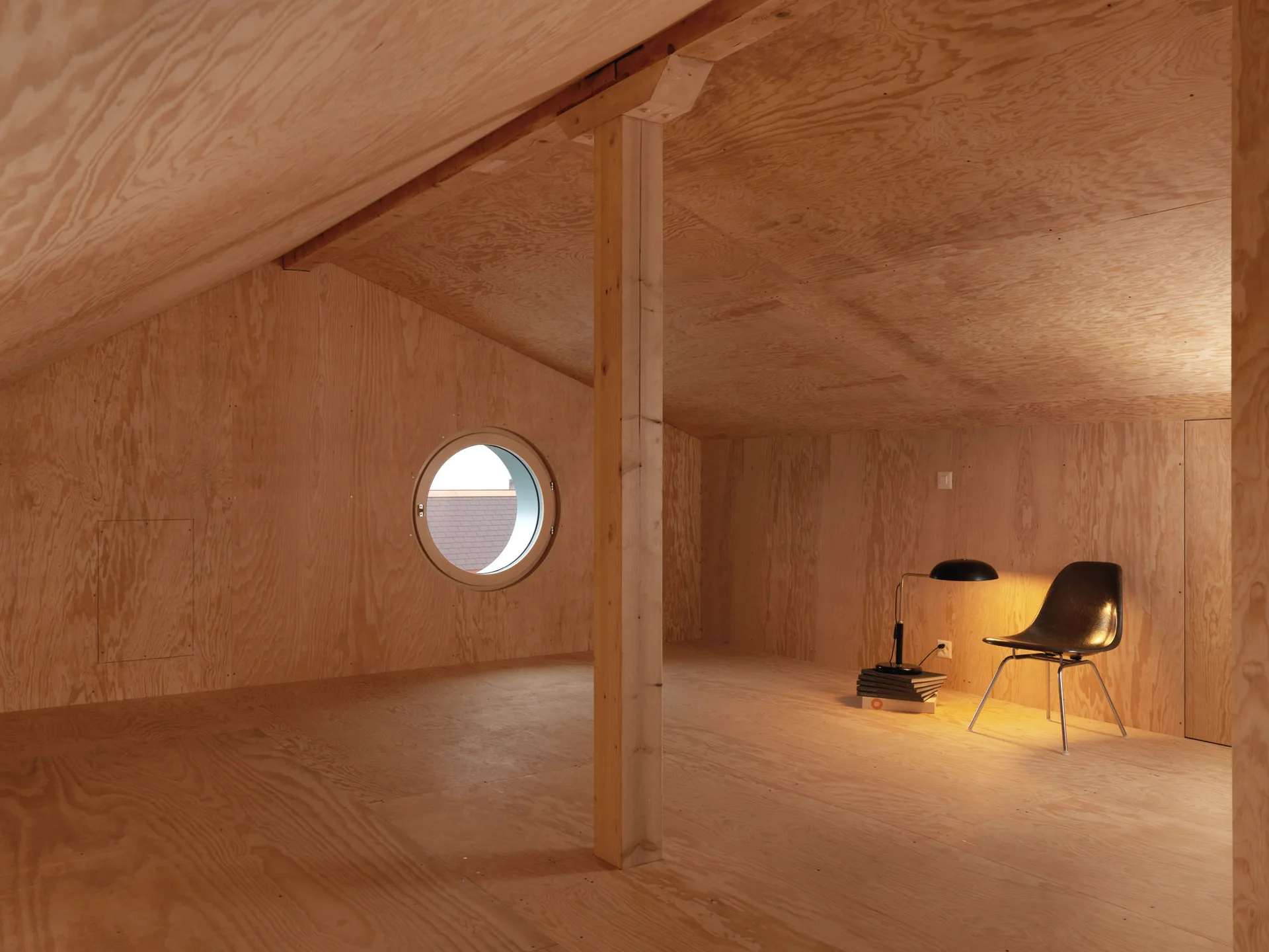 An attic room clad in light wood plywood panels with a circular window, a central column, and a single black chair illuminated by a low lamp.