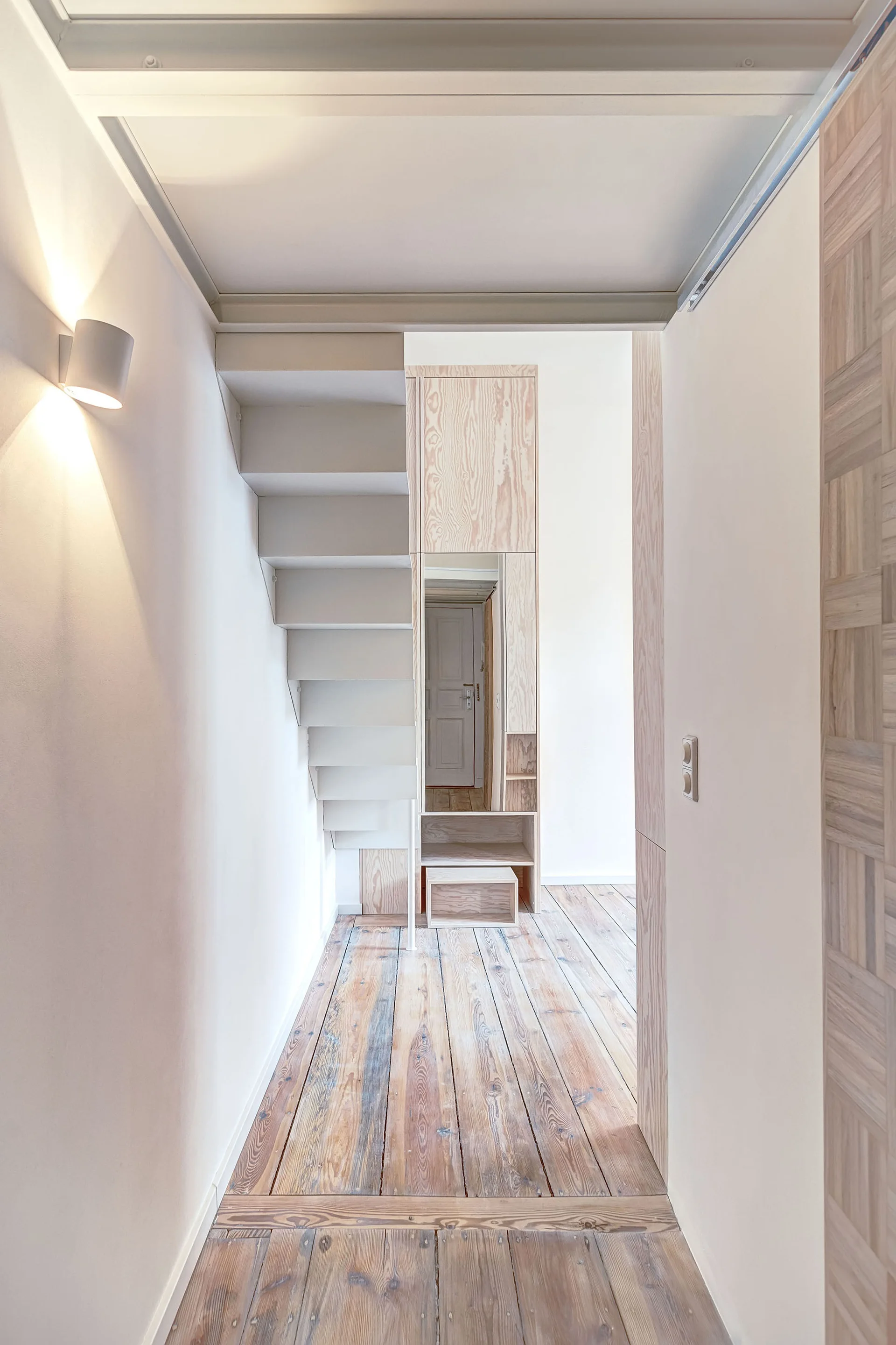 A minimalist hallway features a white structural staircase, distressed wooden floor planks, and a tall light wood built-in cabinet.