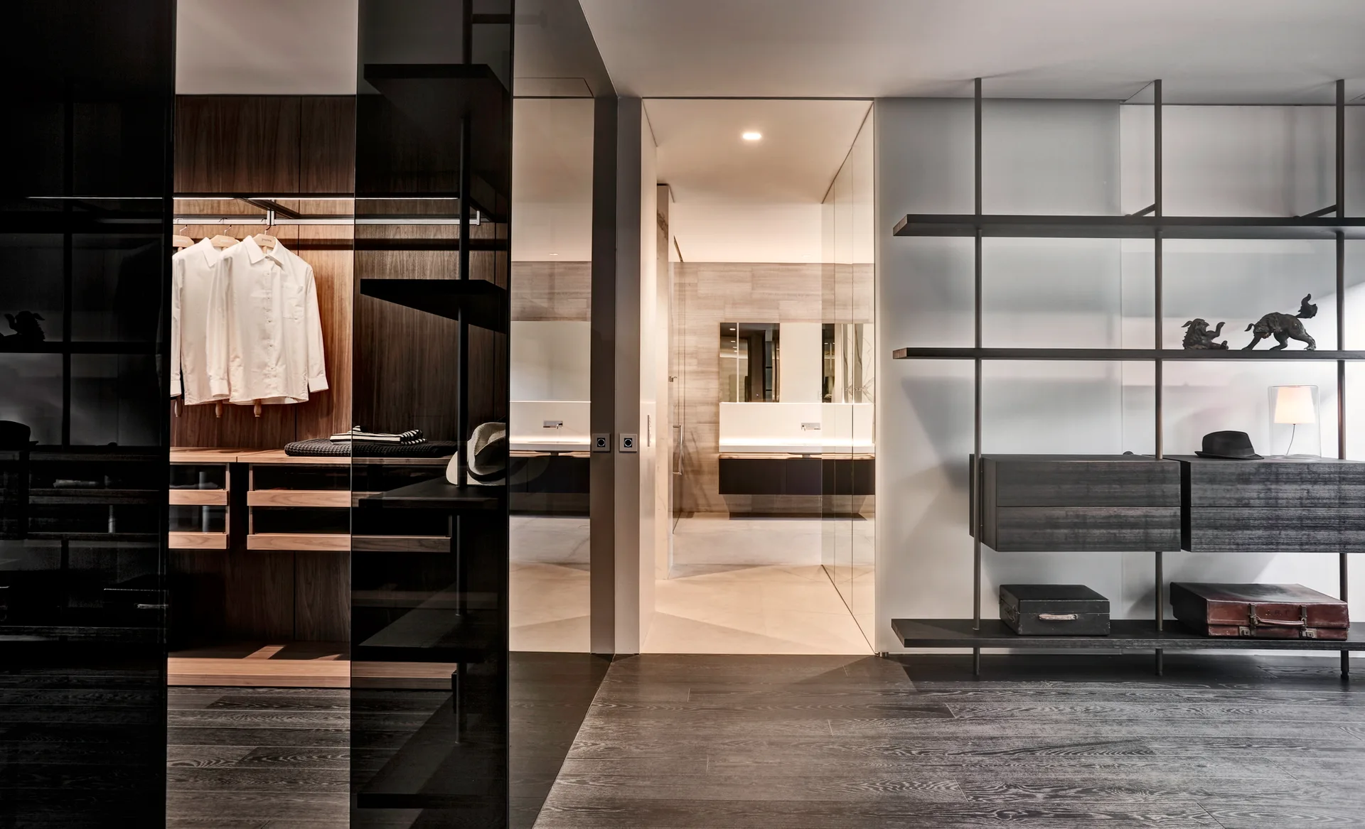 A modern walk-in closet with dark wood built-in wardrobes, a minimalist metal shelving unit, and an open doorway leading to a bright bathroom.