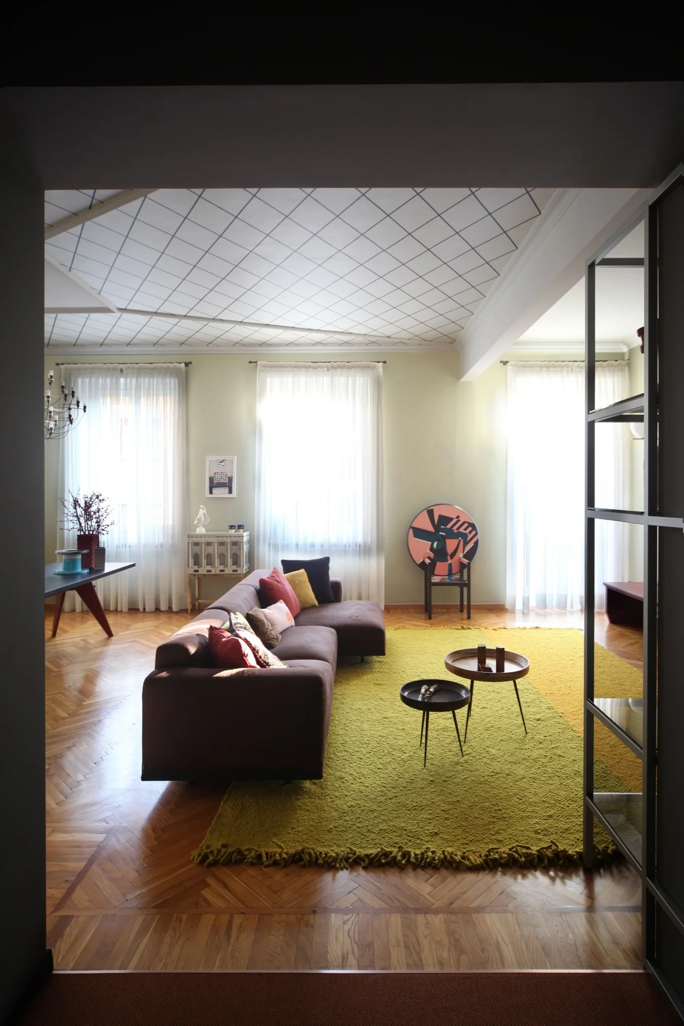 A living room with a brown sectional sofa, a yellow textured rug, herringbone wood floors, and a distinctive white grid-patterned ceiling.