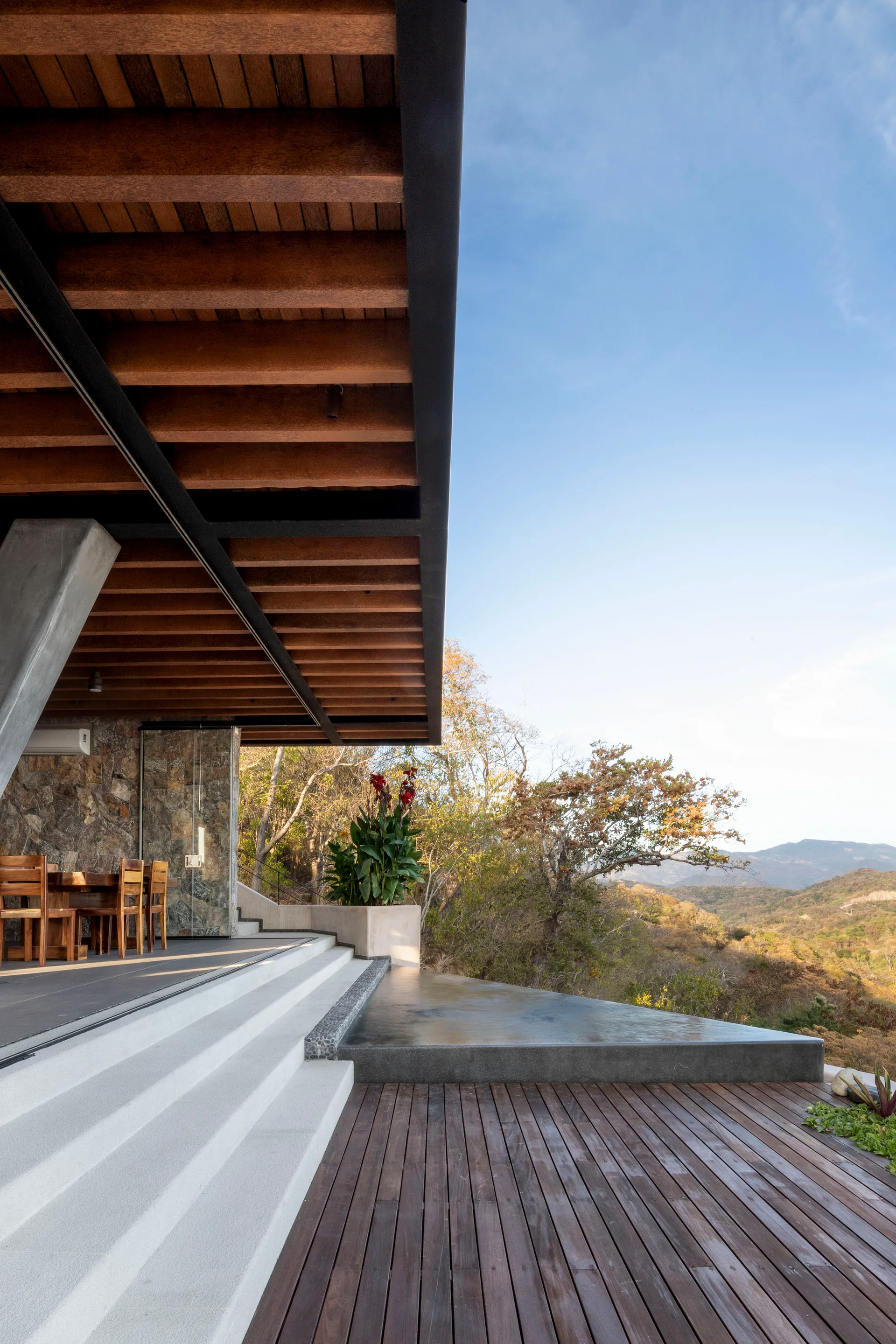 A modern outdoor terrace features a wooden deck, concrete steps leading to a dark infinity pool, and a dining table under a wood-beamed roof.