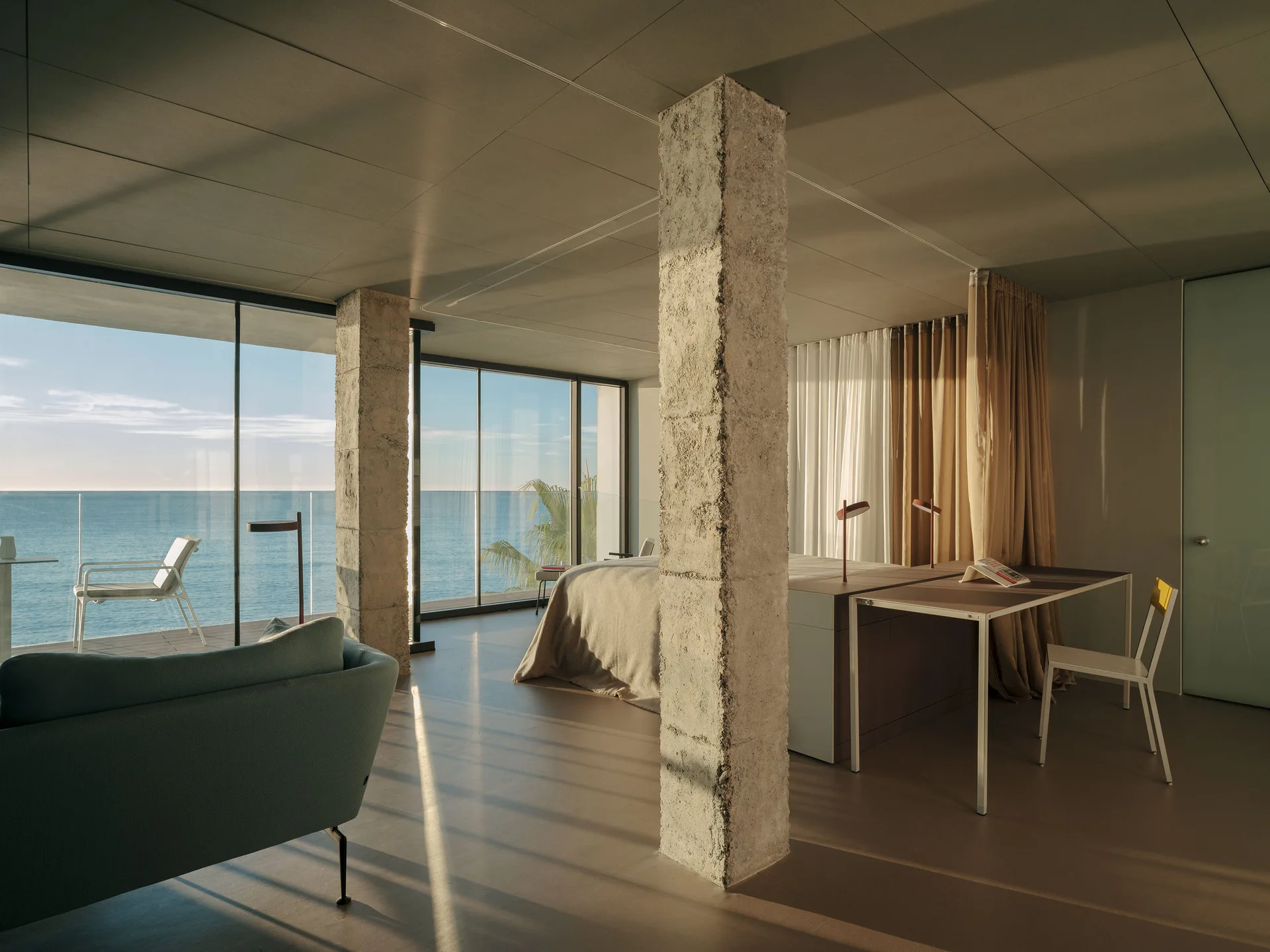 A minimalist bedroom featuring raw concrete pillars, a simple bed and desk, and expansive glass walls overlooking the sea.