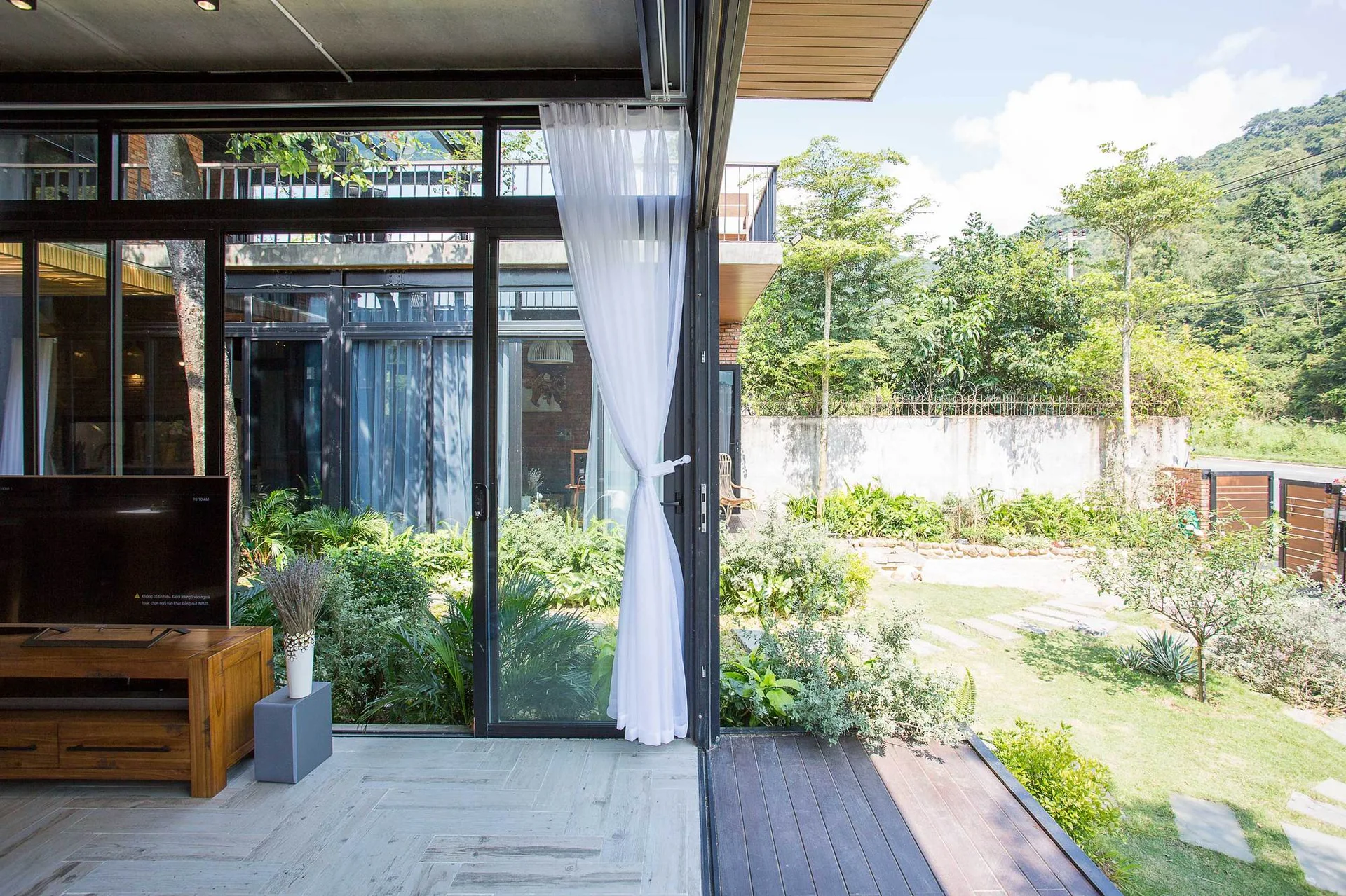A light-filled living room features a wooden media console and large sliding glass doors opening onto a wood-planked deck and lush green backyard.