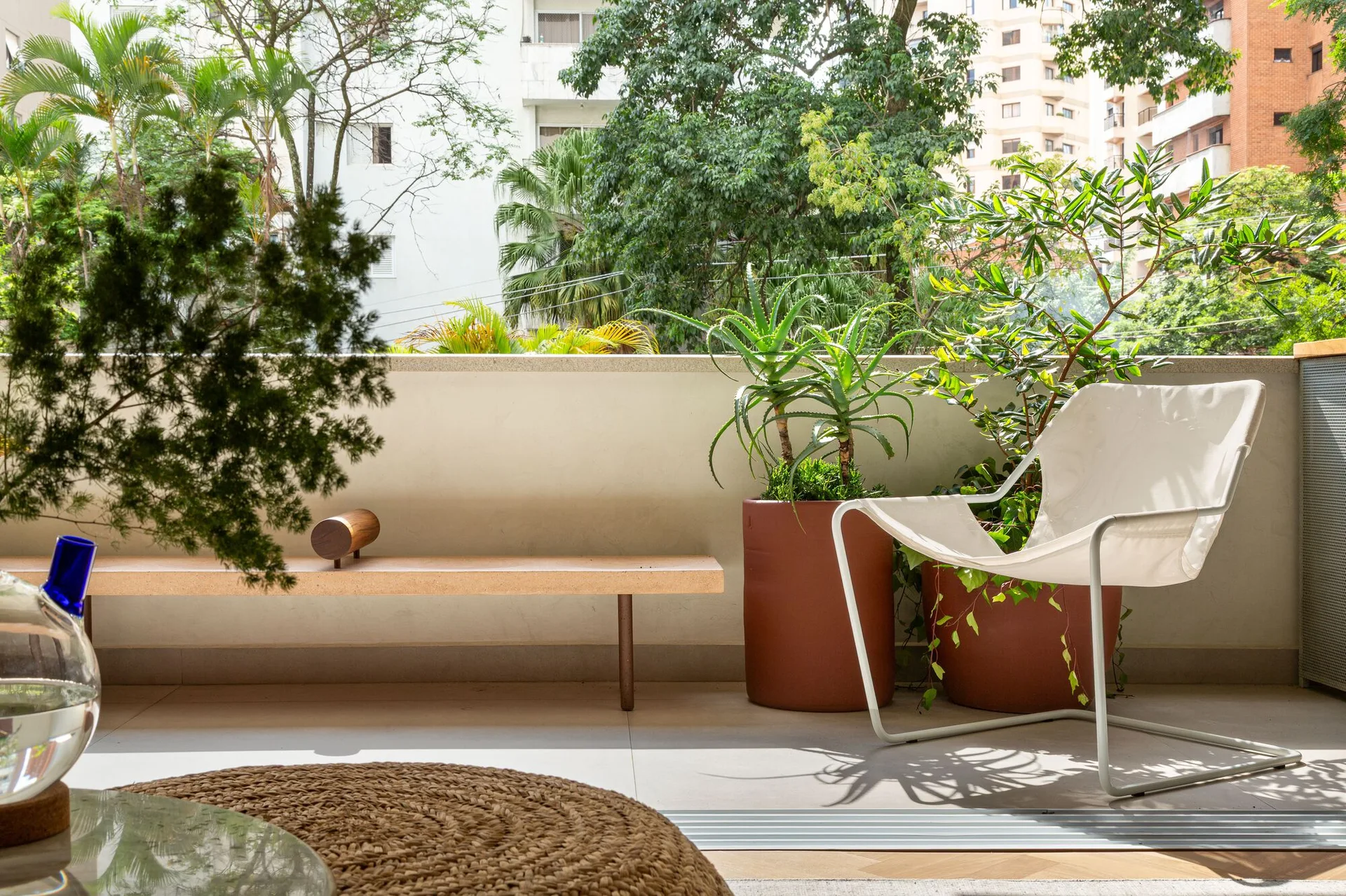 A minimalist outdoor balcony features a white lounge chair, large terracotta planters with green foliage, a low wooden bench, and a woven rug.