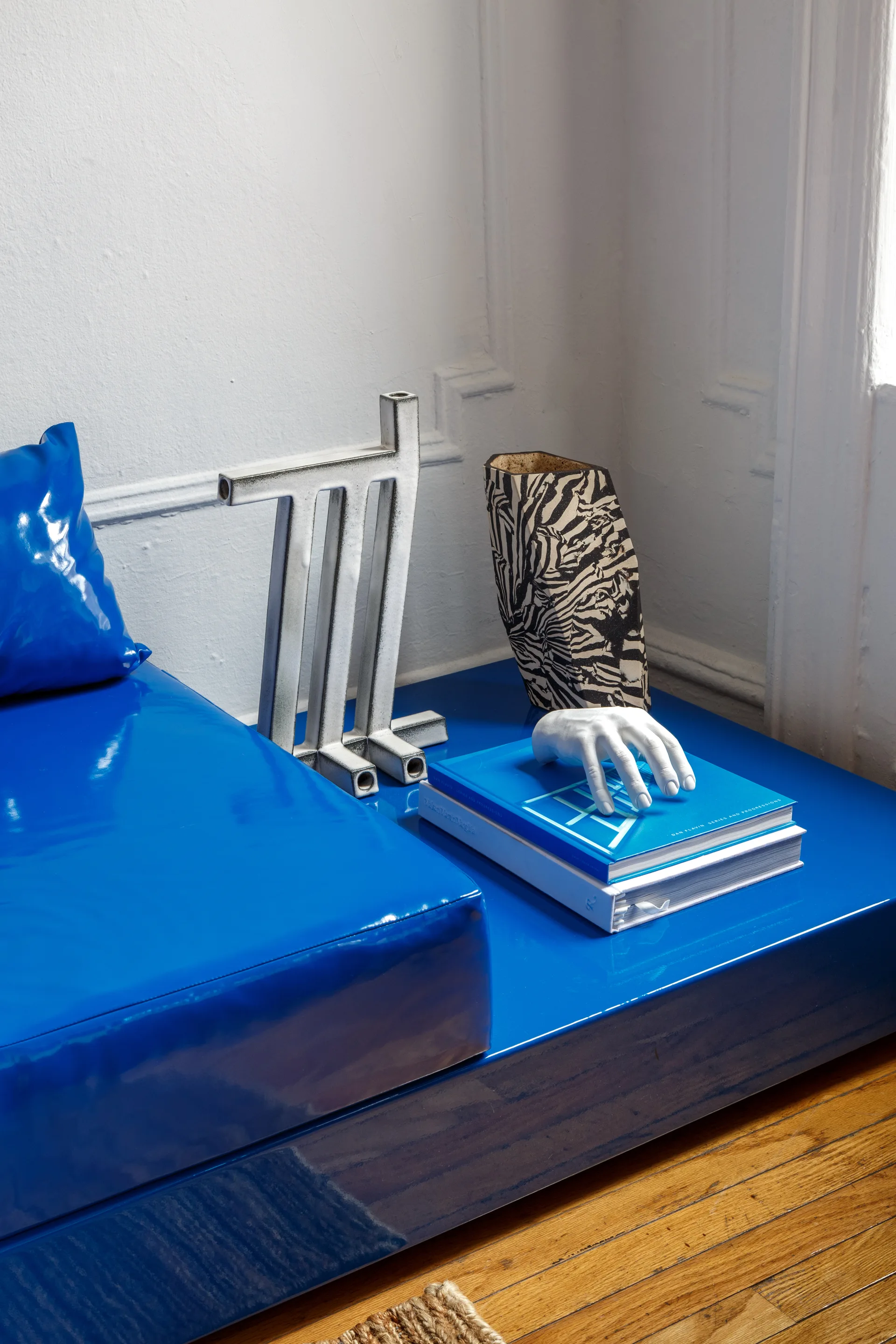 A high-gloss cobalt blue platform bench is decorated with art books, a sculptural white hand, a zebra-patterned vase, and a silver geometric candle holder.