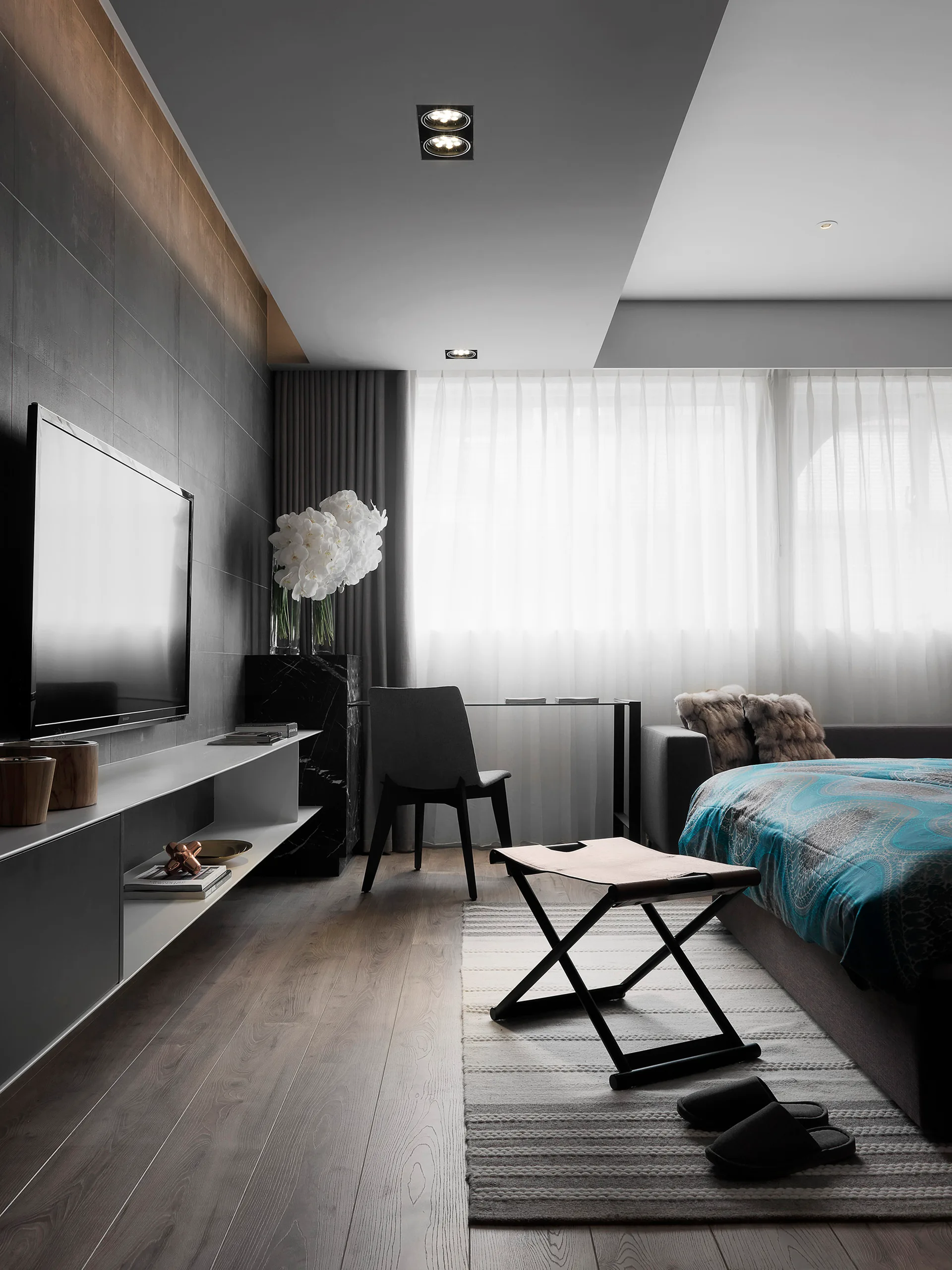 A modern bedroom features a grey paneled wall with a mounted television, a floating white and grey media console, a leather folding stool, and a bed.