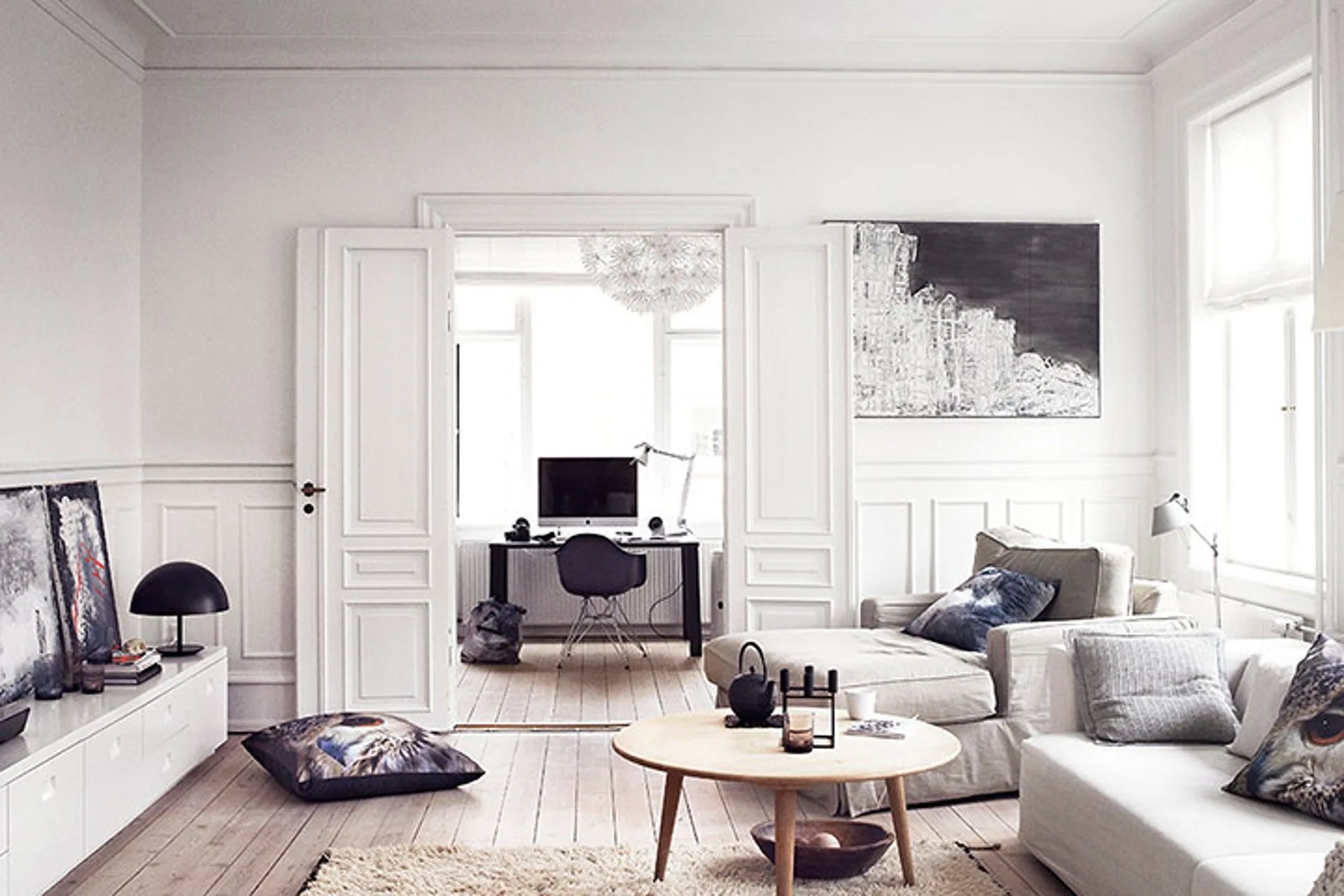 A light-filled living room with white wainscoting, a round wooden coffee table, and open double doors leading into a home office.