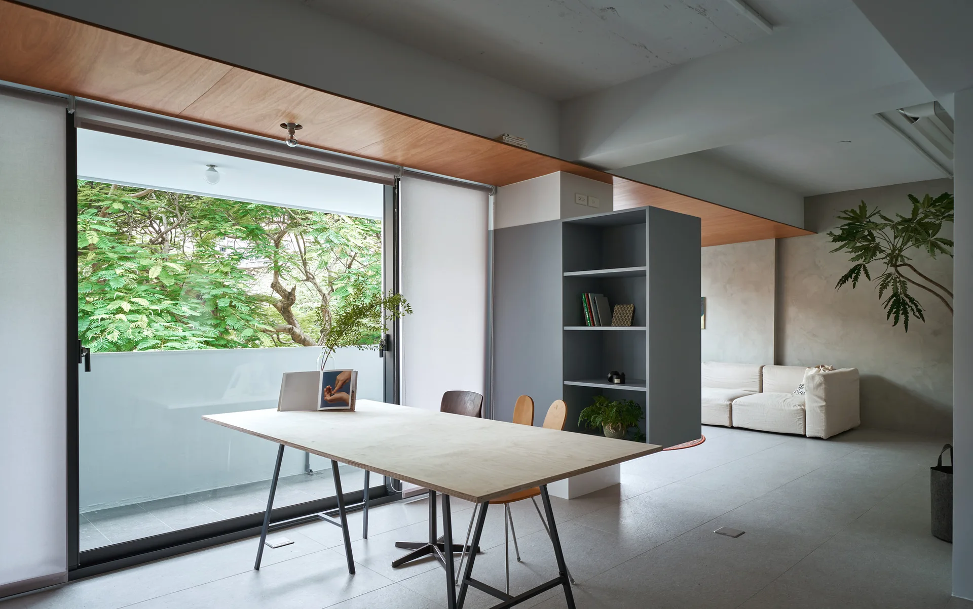 A light wood dining table sits near a sliding glass door next to a gray shelving unit and a concrete-walled living area.