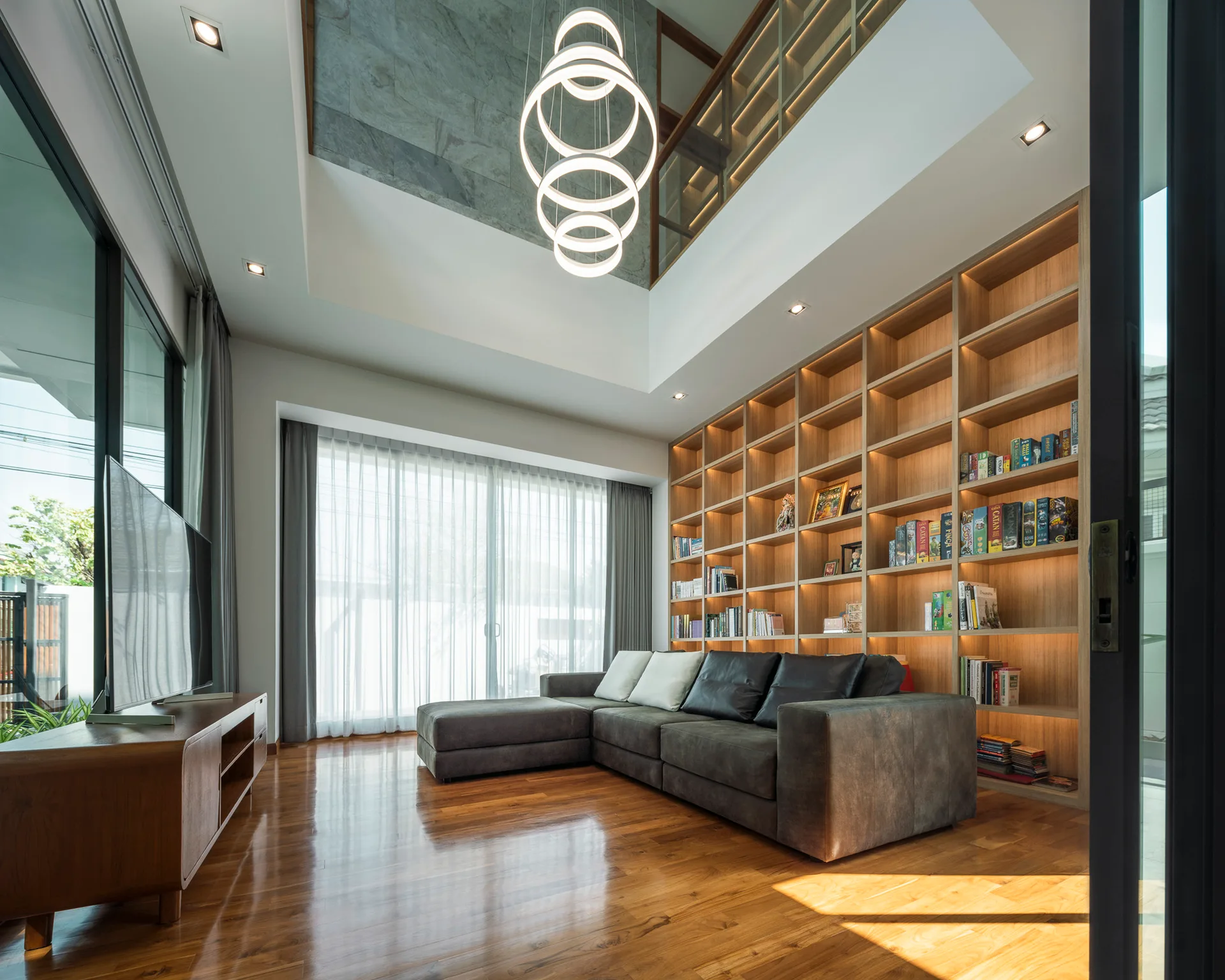 A modern living room with a double-height ceiling, a large built-in wooden bookcase with integrated lighting, and a dark grey sectional.