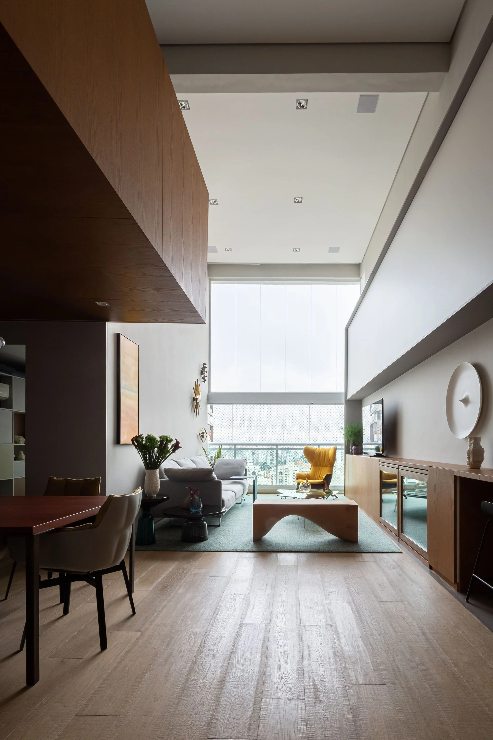A double-height living room featuring a grey sofa, a curved wooden coffee table, a yellow armchair, and large windows.
