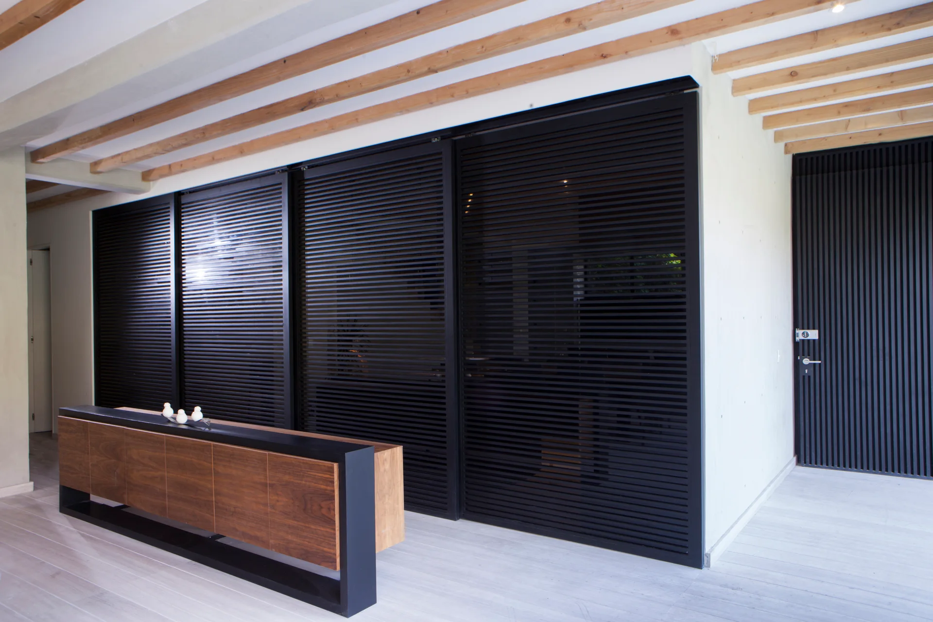 A modern foyer features a wood console with a black frame, large black slatted sliding doors, and exposed natural wood ceiling beams.