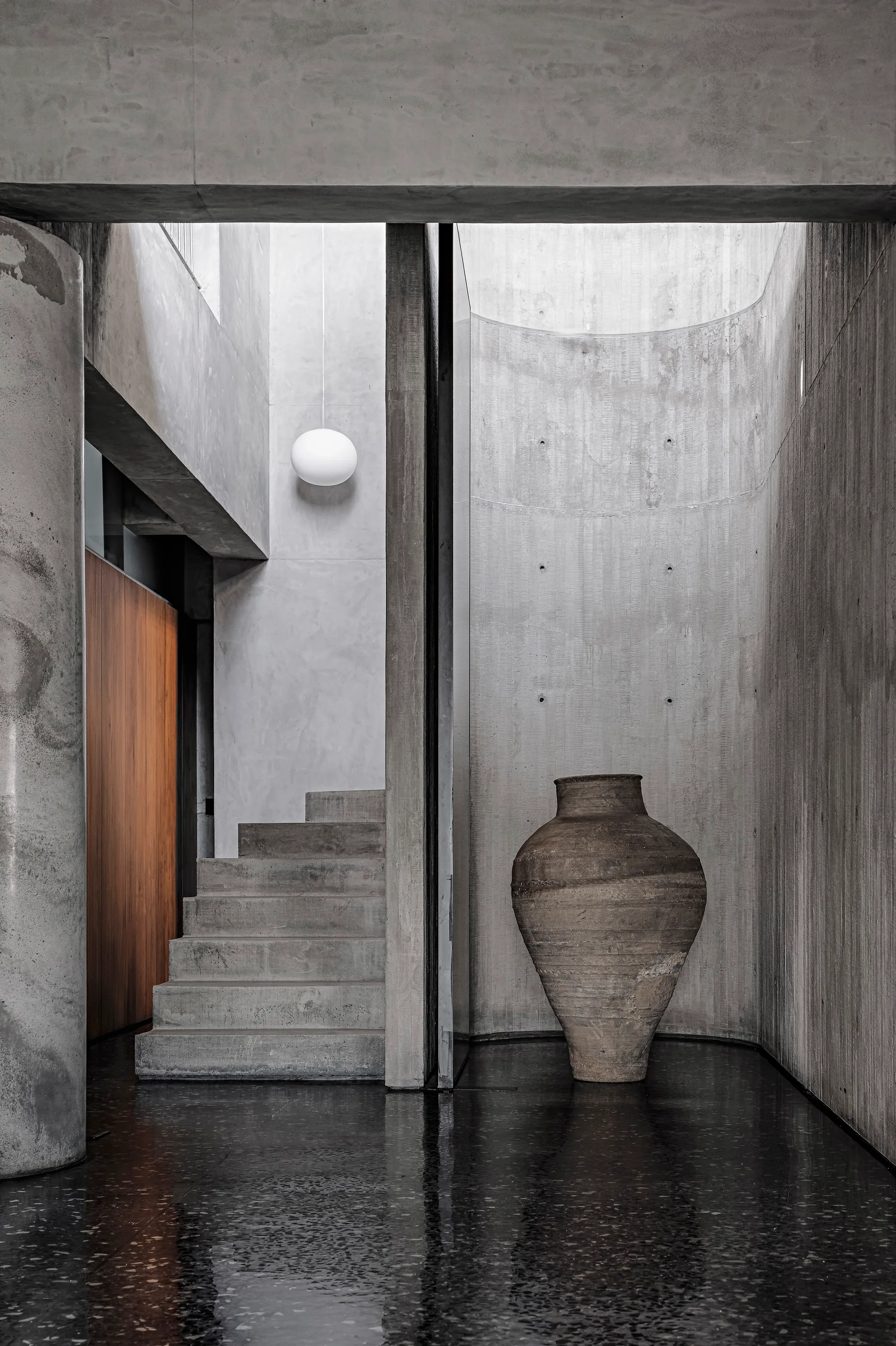 A raw concrete entryway features a concrete staircase, a large weathered clay vessel in a curved alcove, and a dark polished floor.