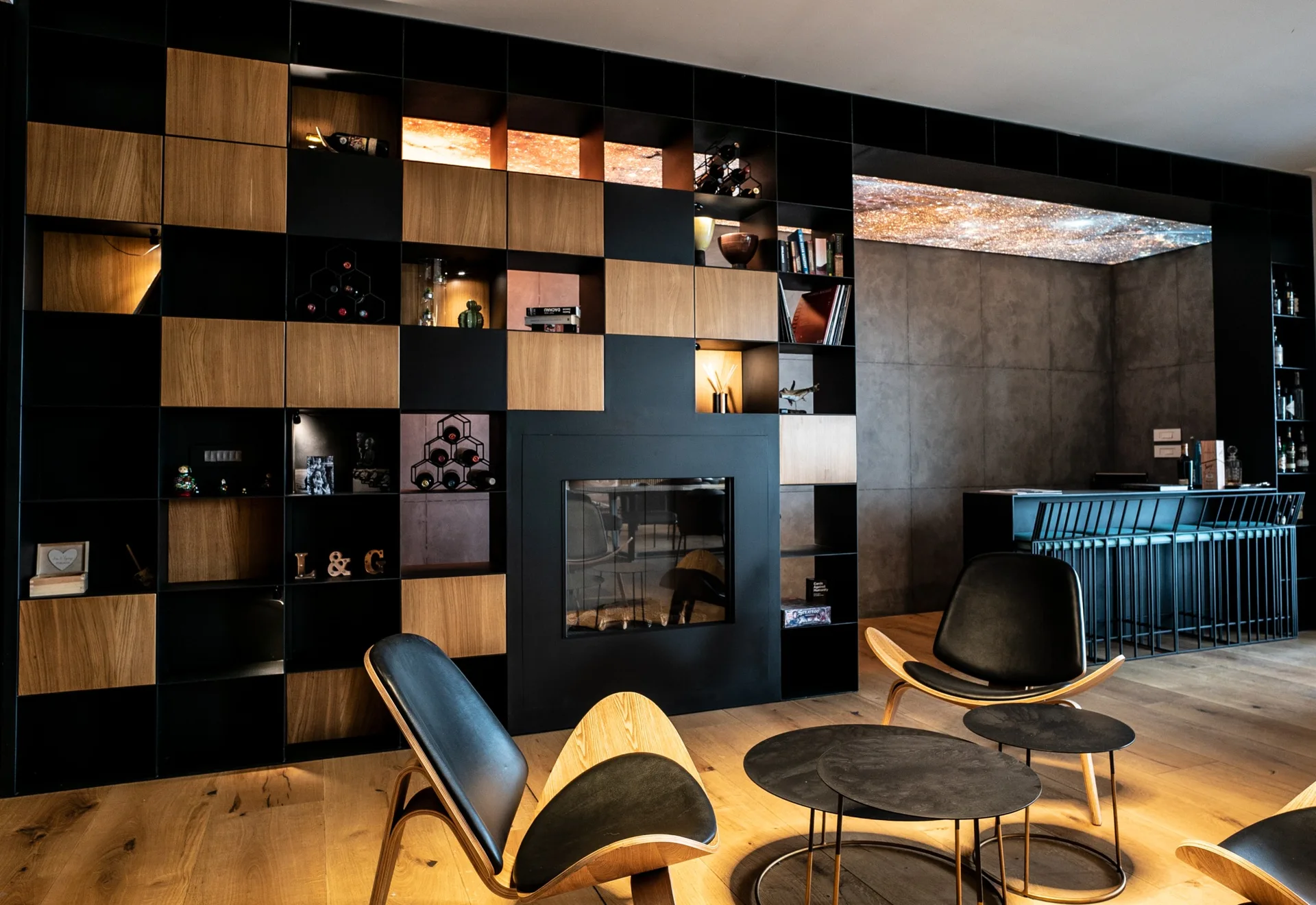 A contemporary living room features a black and wood grid wall unit with an integrated fireplace, molded plywood lounge chairs, and a bar area.
