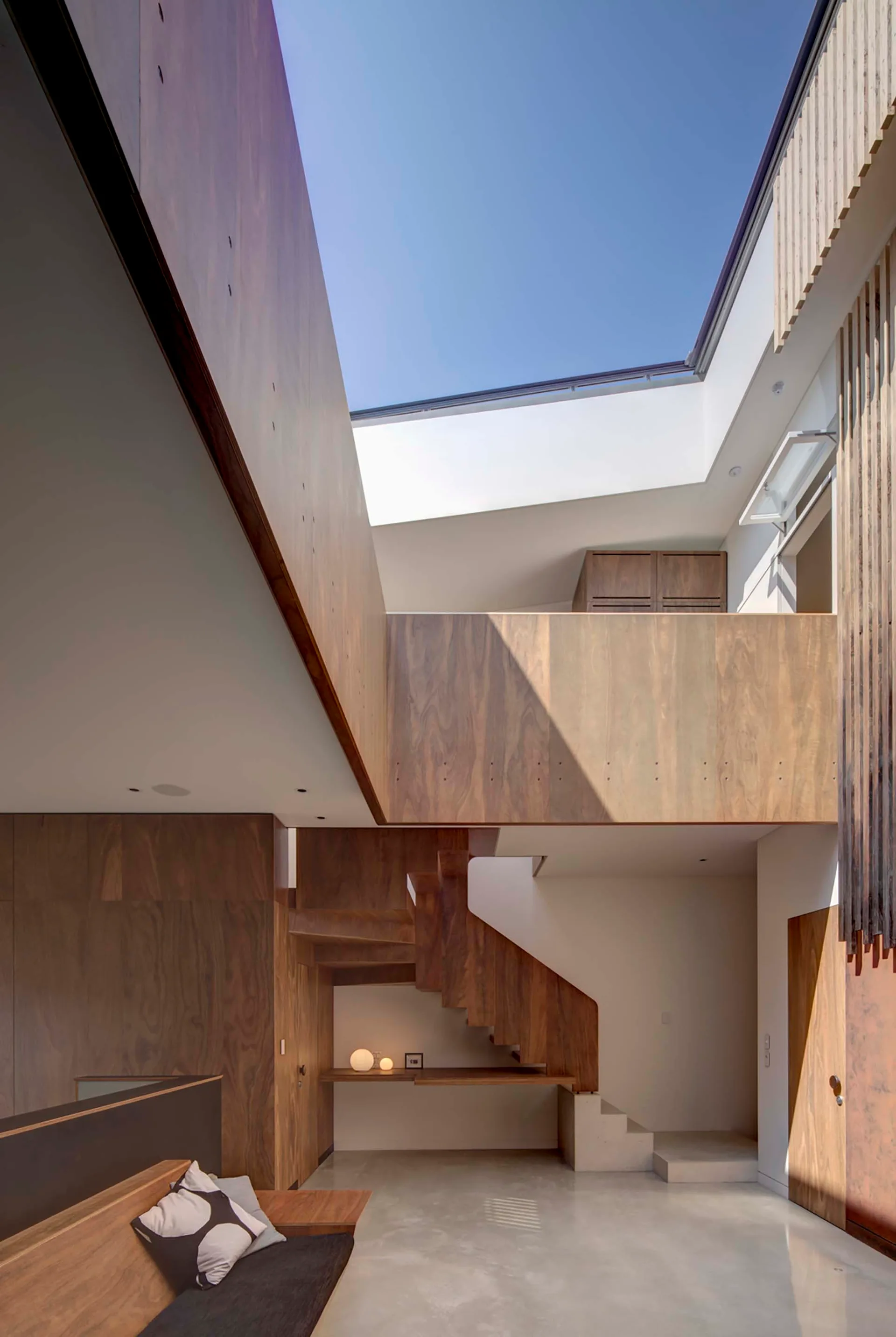 A contemporary interior featuring a sculptural wooden staircase and extensive wood paneling underneath a large rectangular skylight showing a blue sky.