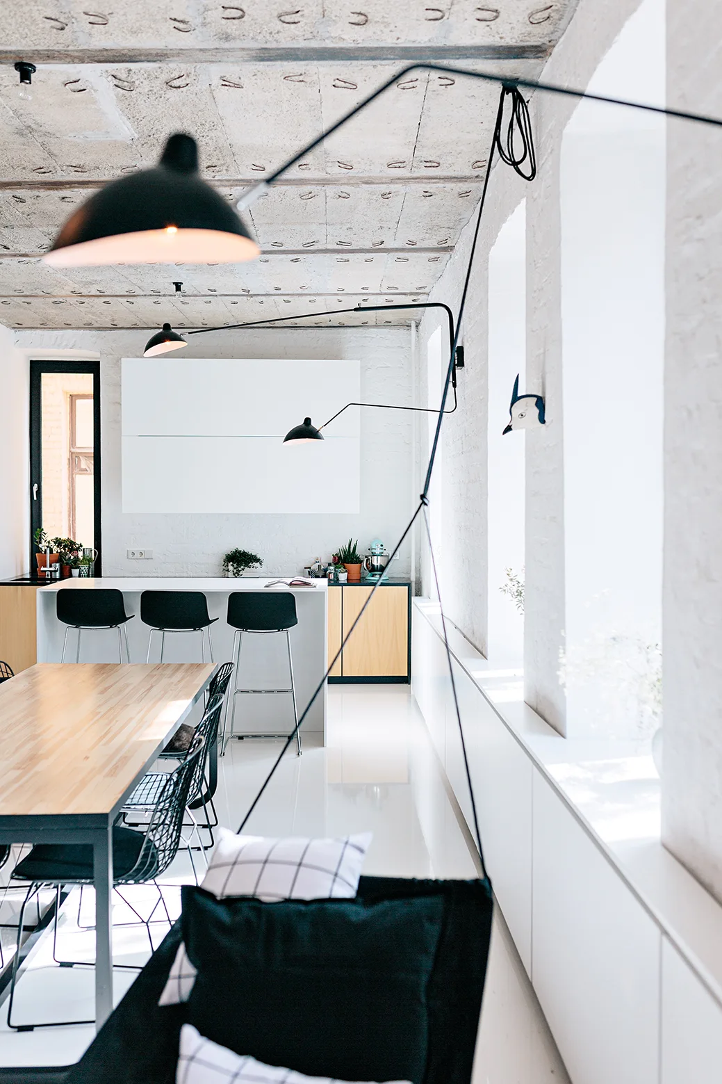 An open-concept kitchen features a wood dining table, a white island with bar stools, black articulating lamps, and an exposed concrete ceiling.
