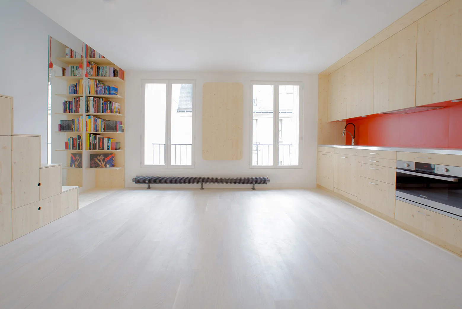 A minimalist room features light wood kitchen cabinetry with a bright orange backsplash, adjacent to two windows and a tall bookshelf.