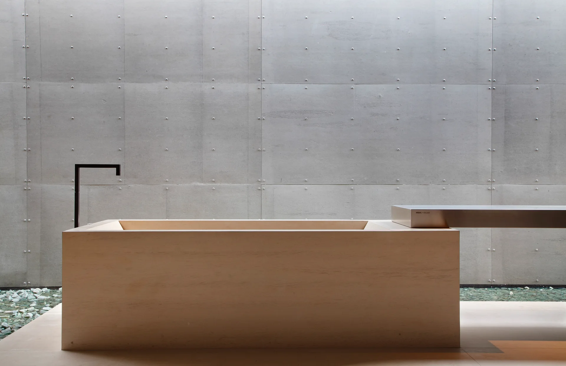 A minimalist rectangular light wood bathtub sits against a grey concrete paneled wall with a black angular wall-mounted faucet.