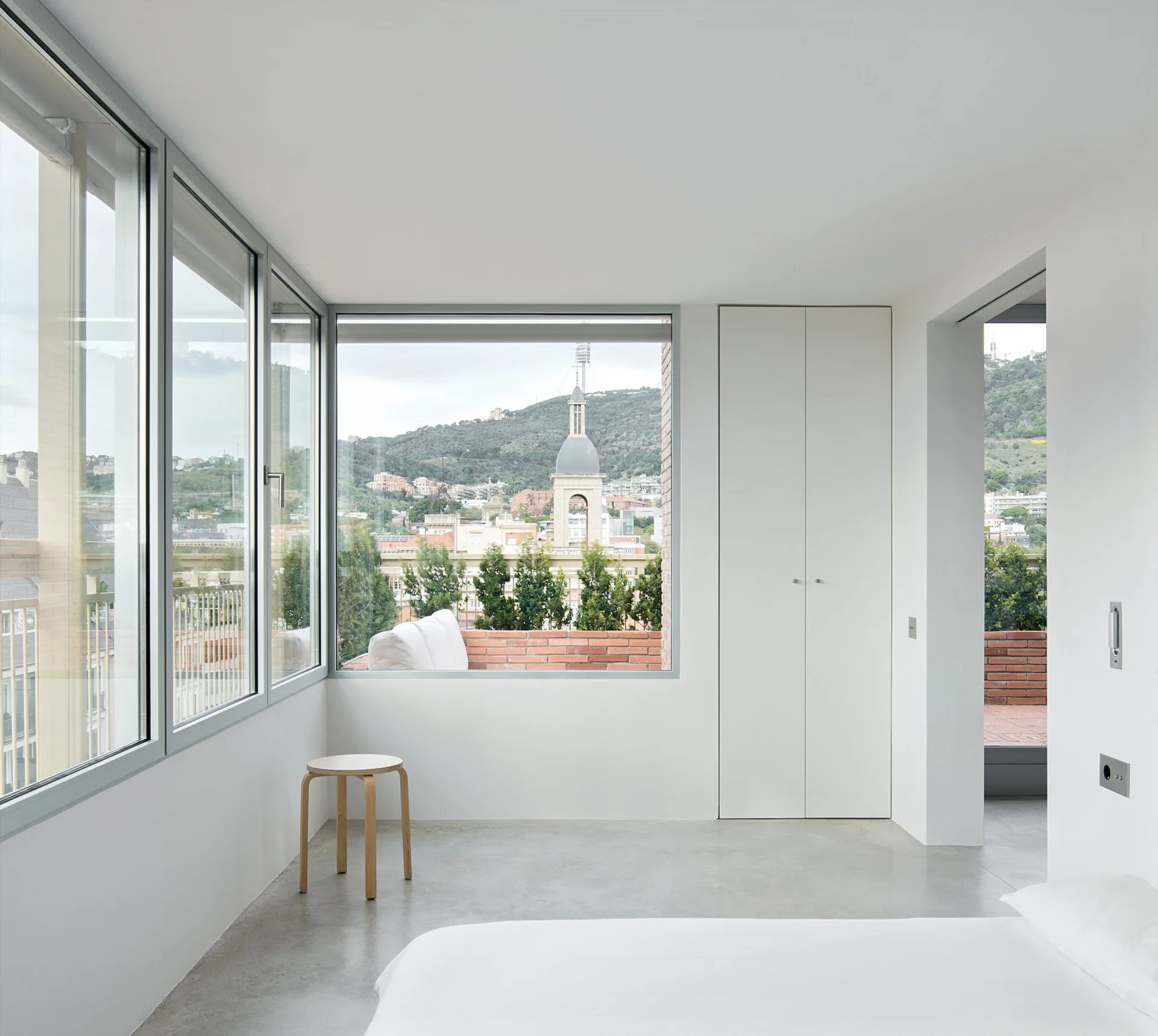 A minimalist bedroom with a grey concrete floor, a white built-in wardrobe, and large corner windows showing an urban view.