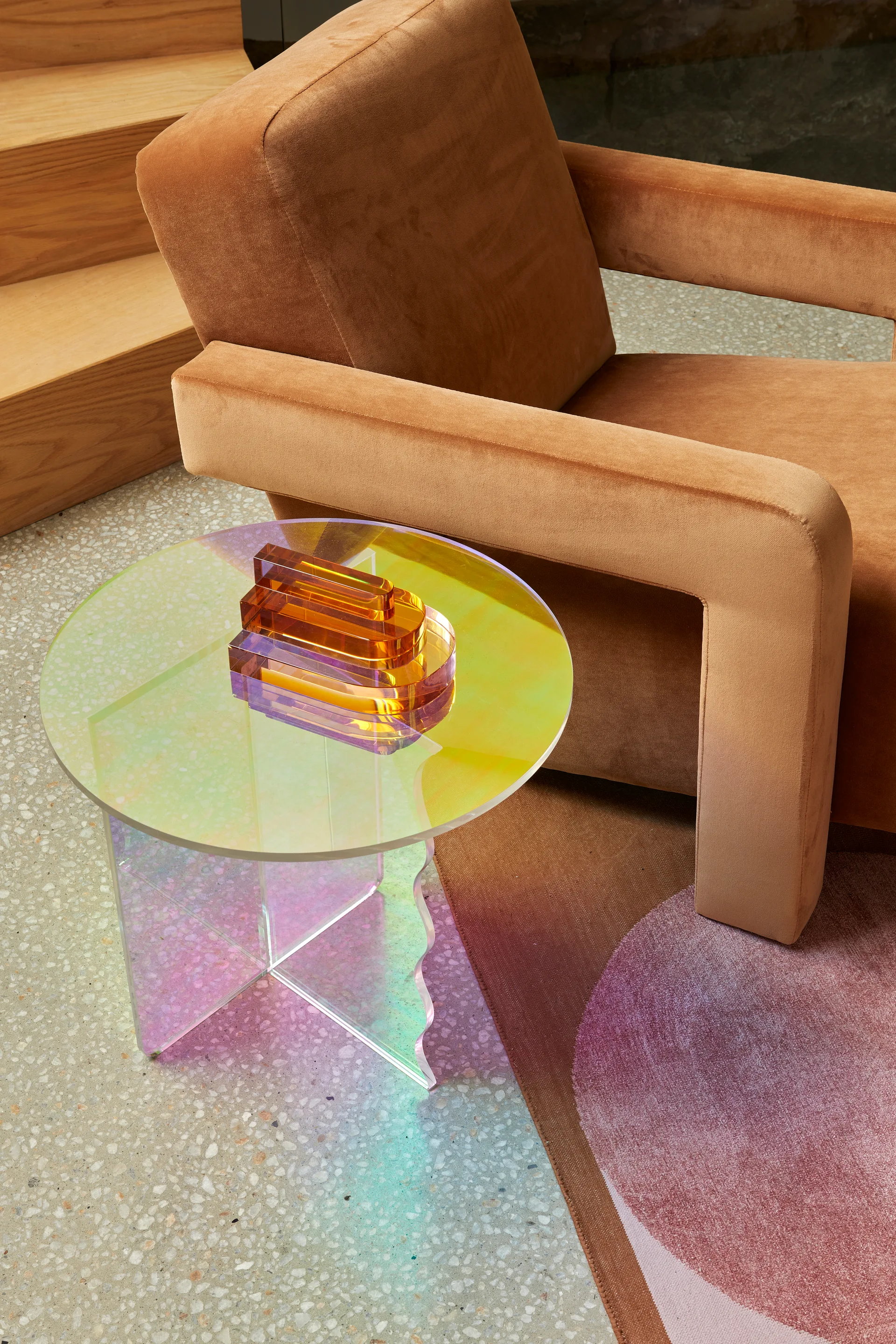 A warm brown upholstered armchair sits next to a circular iridescent side table topped with an amber glass sculpture on a speckled terrazzo floor.