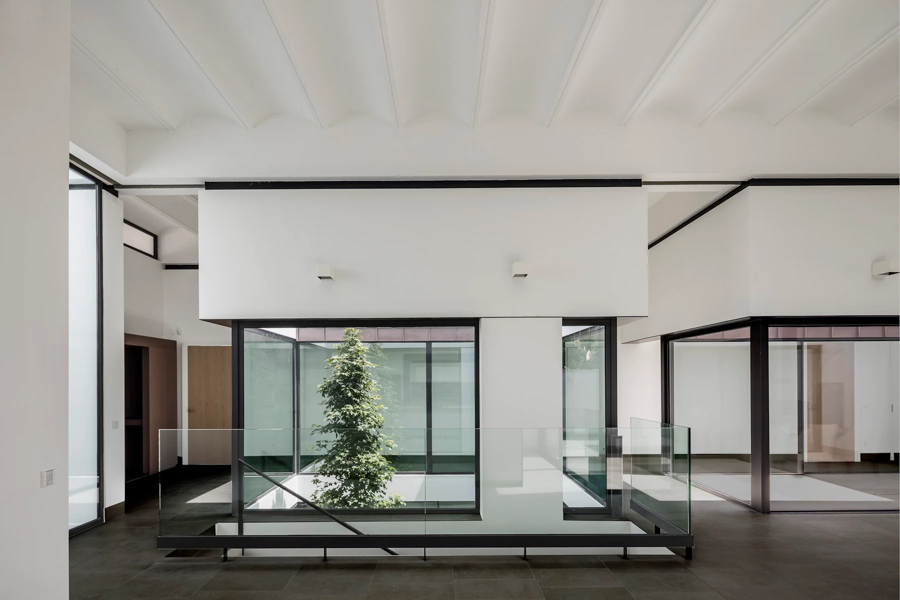 A minimalist hallway landing featuring dark grey tiled floors, a glass balustrade, and large windows looking into an enclosed courtyard with a green tree.