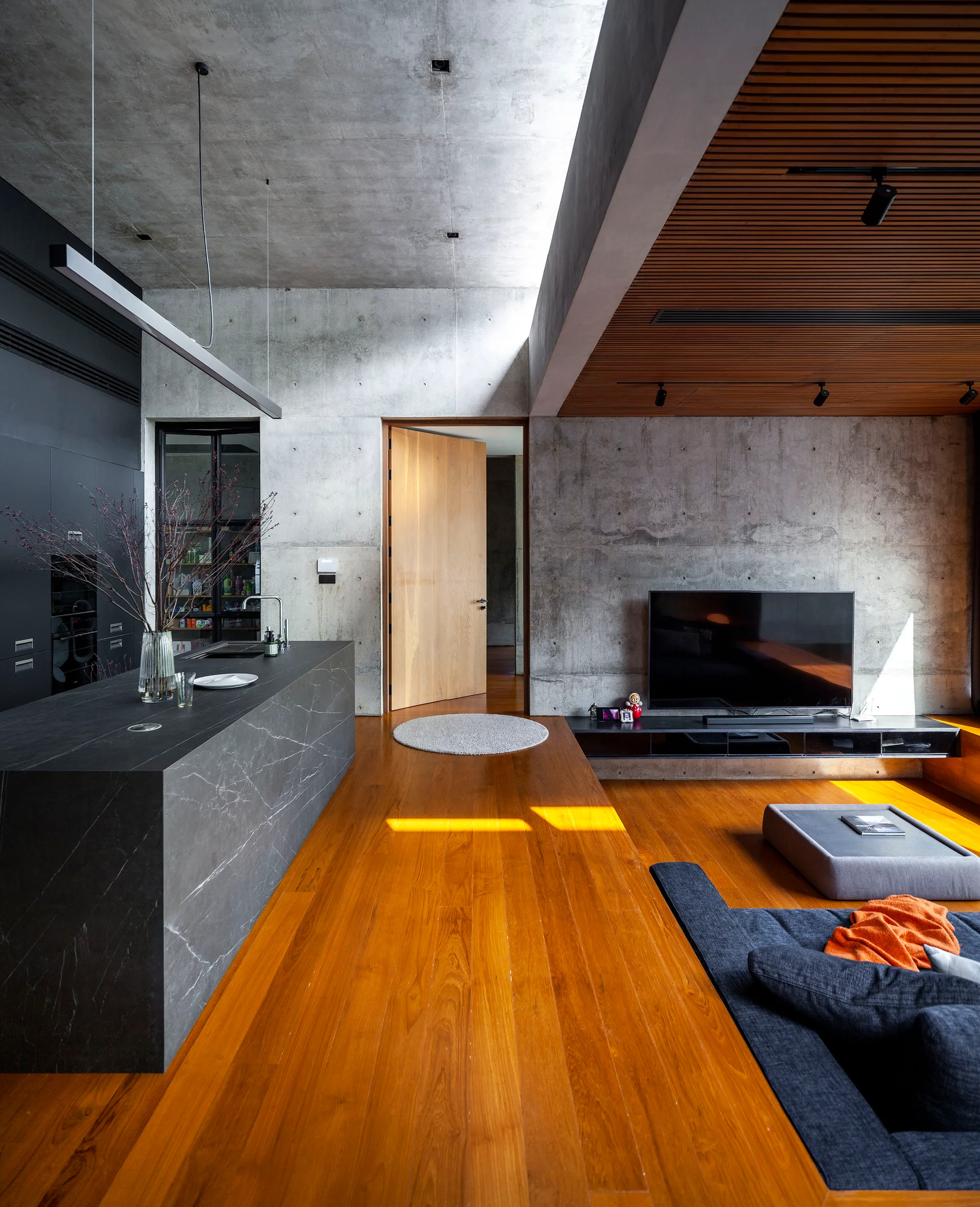 A modern open-concept living space with exposed concrete walls, a dark kitchen island, warm wood flooring, and a slatted timber ceiling panel.