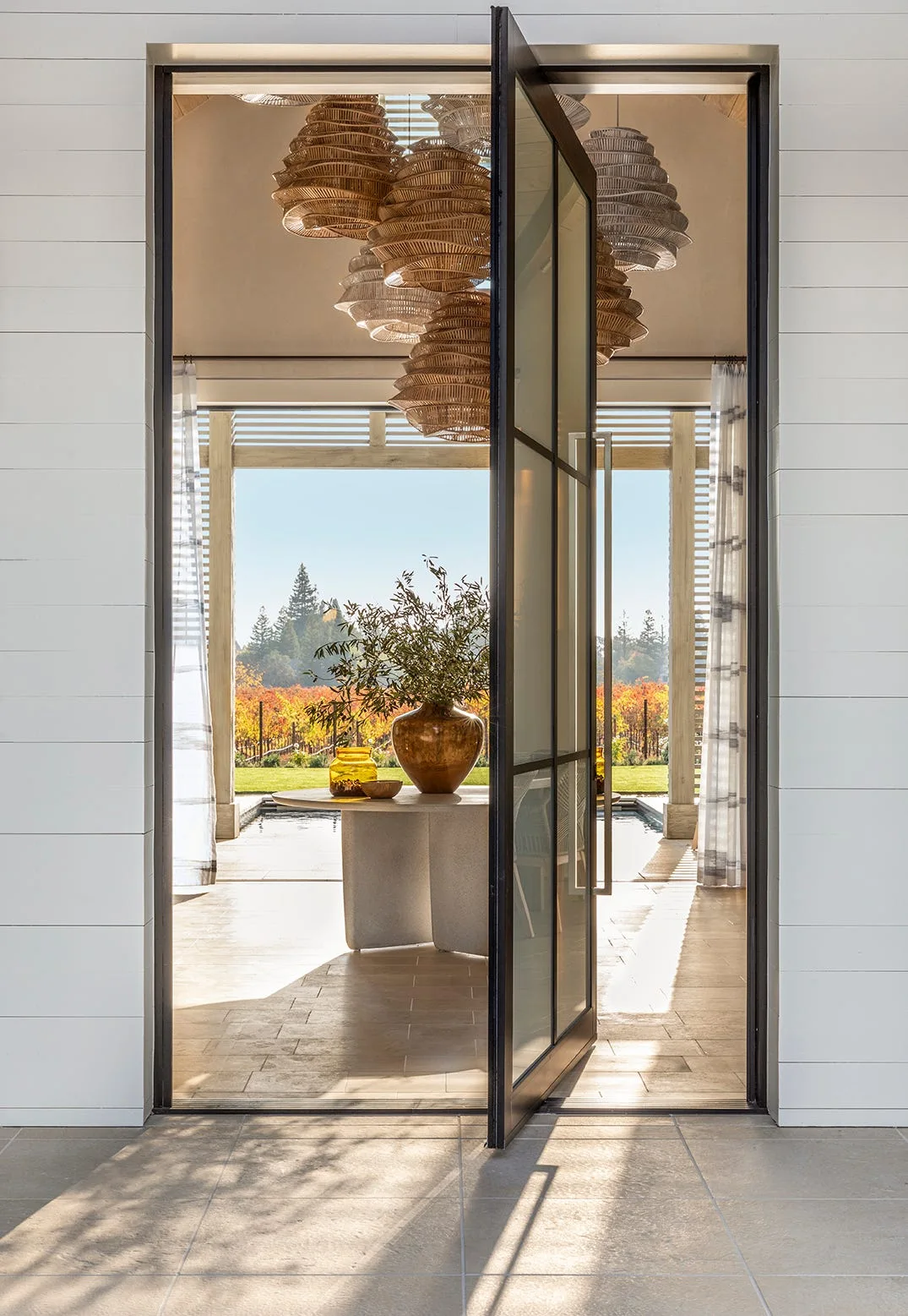 An open black-framed glass pivot door reveals an indoor-outdoor entryway featuring a stone pedestal table and several hanging woven pendant lights.