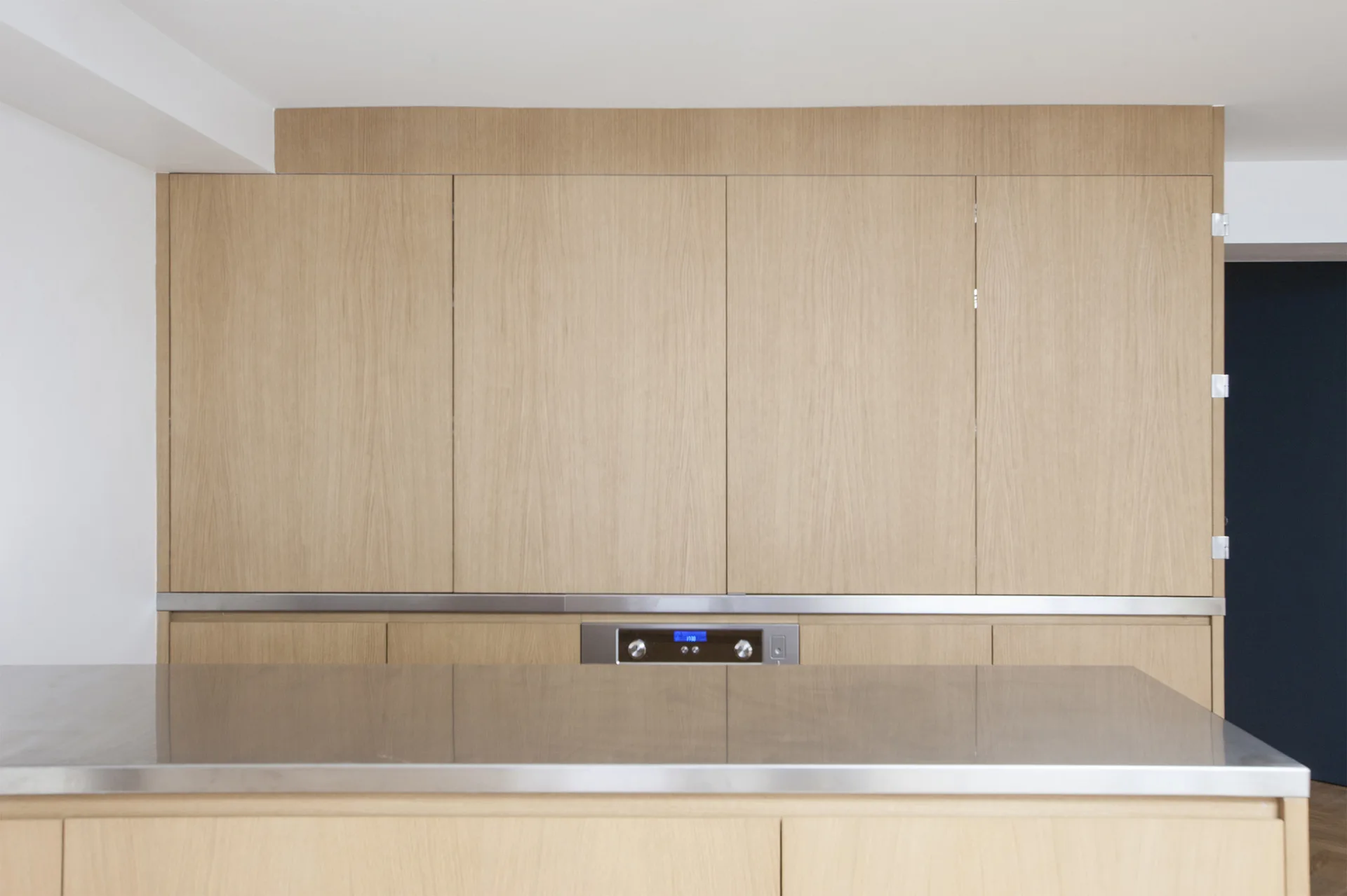 A minimalist kitchen features light wood flat-panel cabinets, an integrated oven, and a stainless steel island countertop in the foreground.