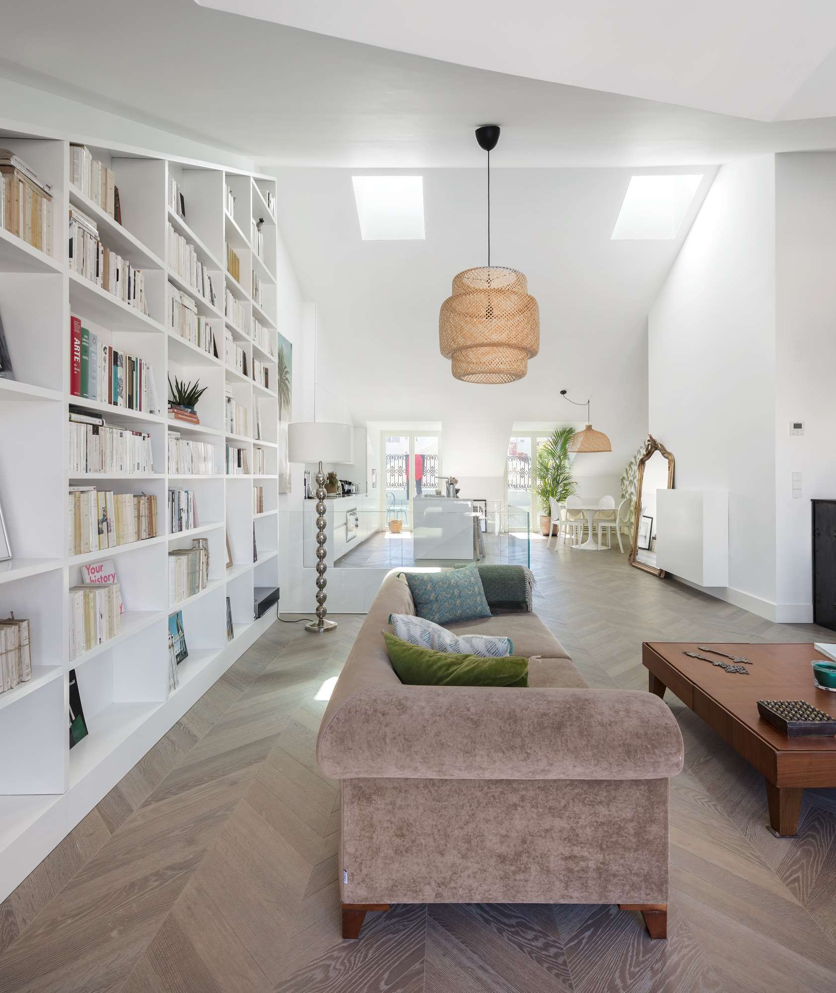 An open living room features a tall white built-in bookshelf, herringbone wood floors, a tan sofa, and skylights in a vaulted ceiling.
