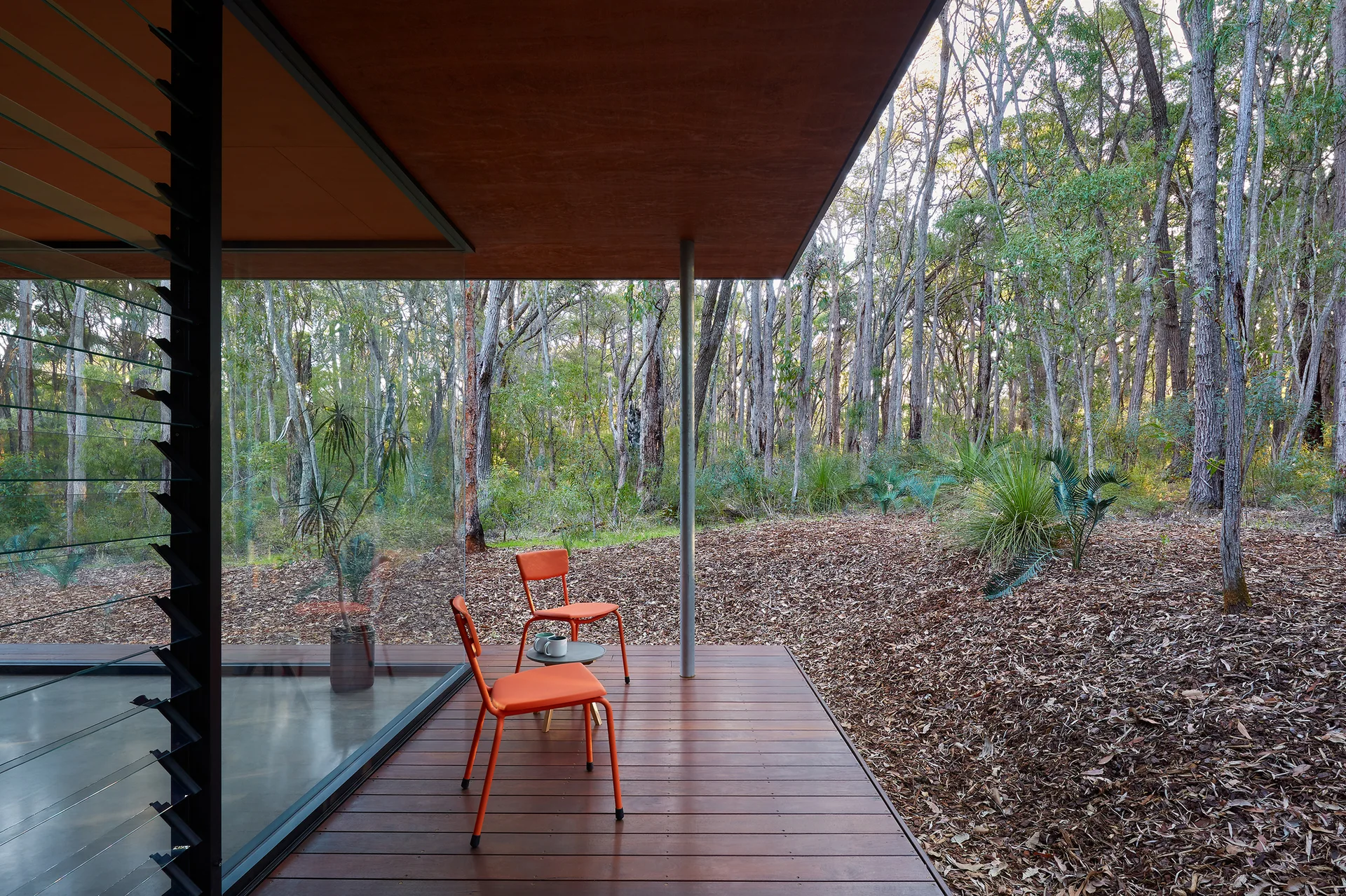 A covered wooden deck with two orange chairs and a small table looks out onto a dense forest through a floor-to-ceiling glass wall.