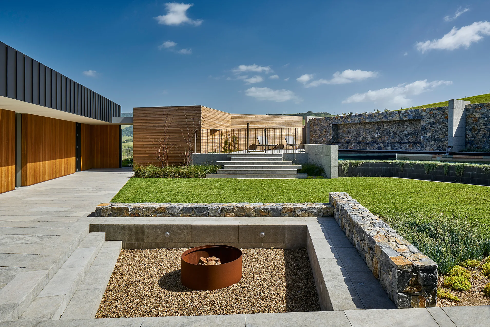 A modern outdoor courtyard features a sunken gravel fire pit area next to a manicured green lawn, stone steps, and timber-clad walls.