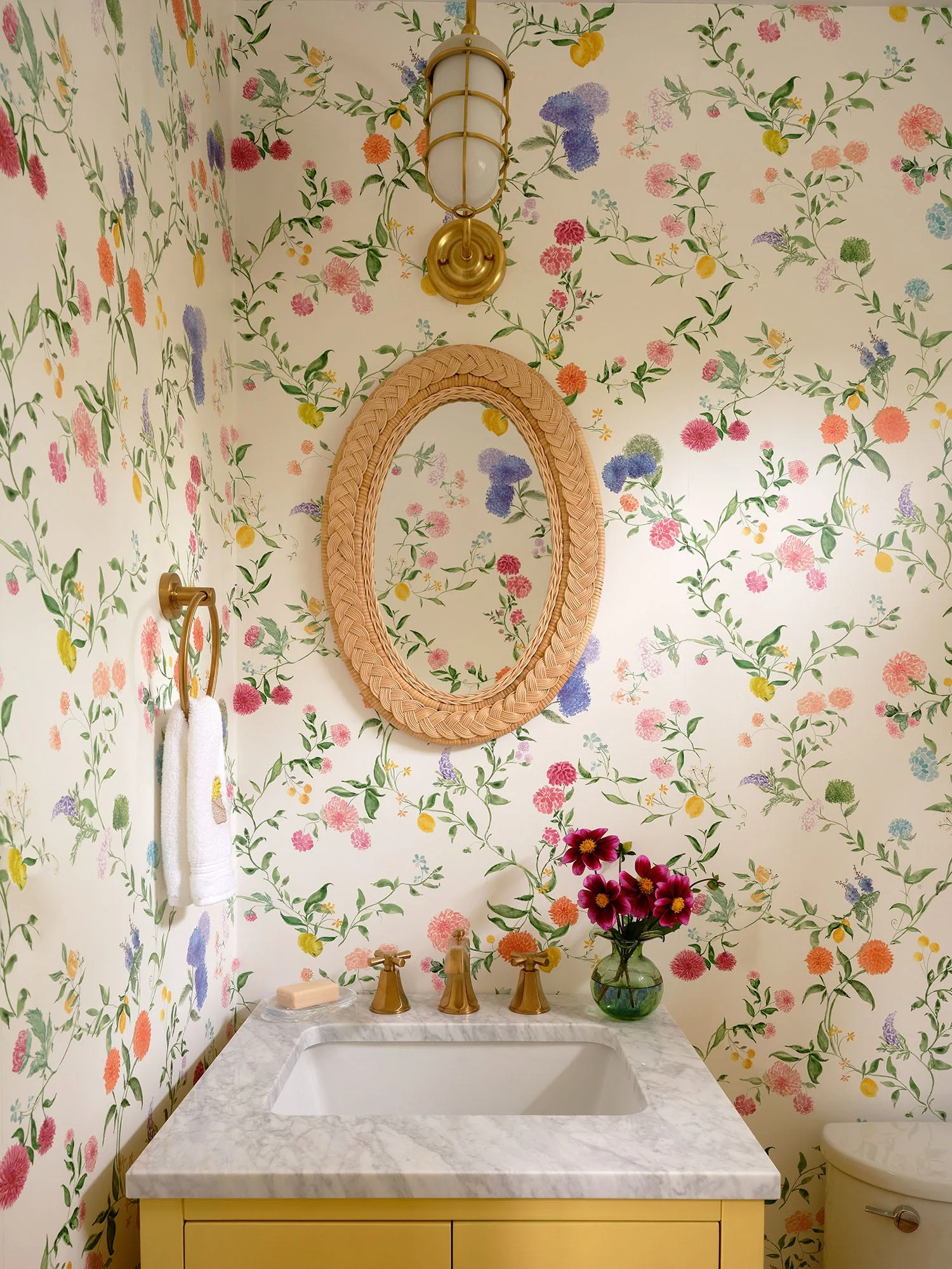 A powder room features botanical floral wallpaper, an oval rattan mirror, a brass wall sconce, and a yellow vanity with a marble countertop.