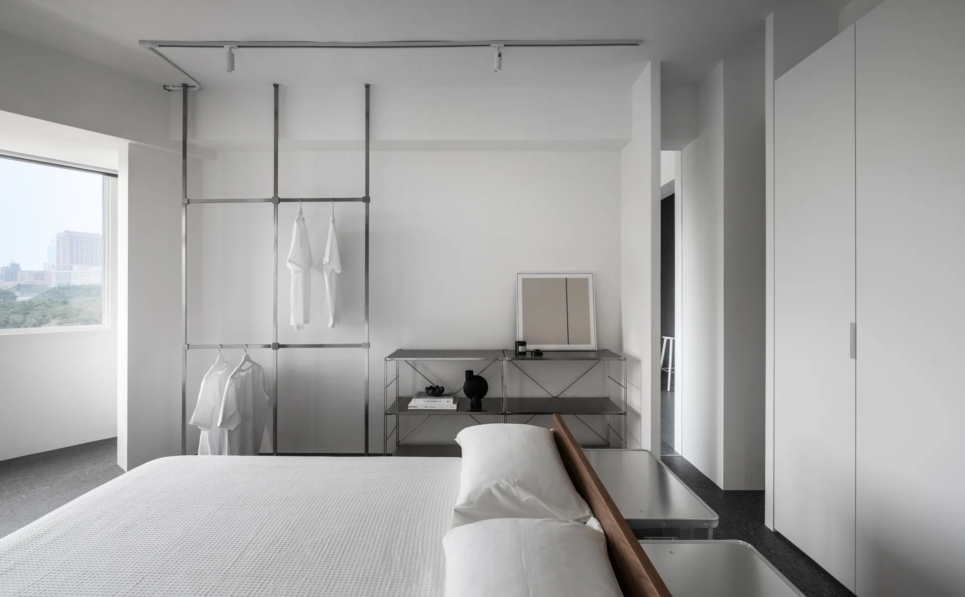 A minimalist white bedroom with a bed in the foreground, a tall metal clothing rack, and integrated white wardrobes against a dark speckled floor.