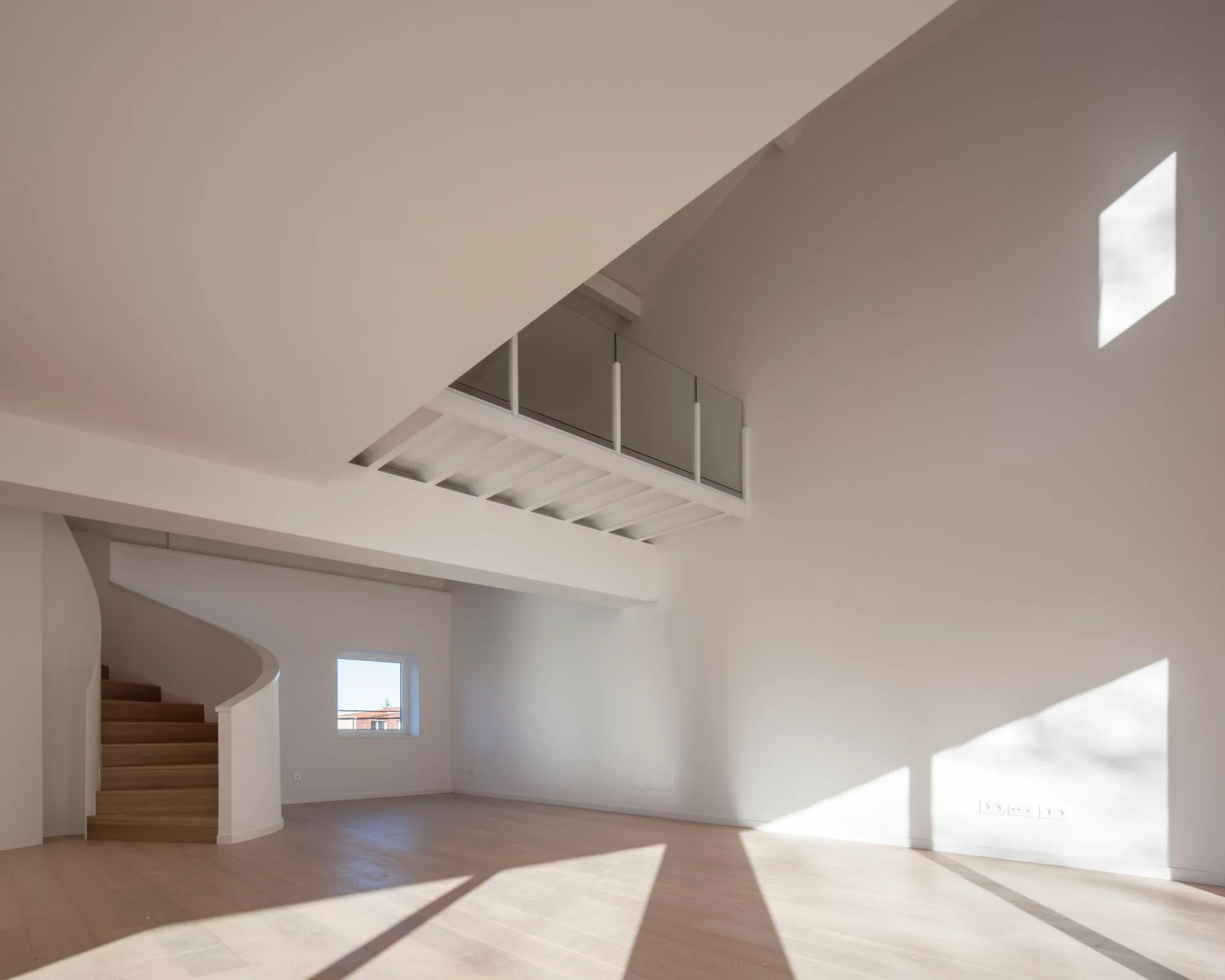 A minimalist white room features a curved wooden staircase and a mezzanine level with a glass railing under a high ceiling.