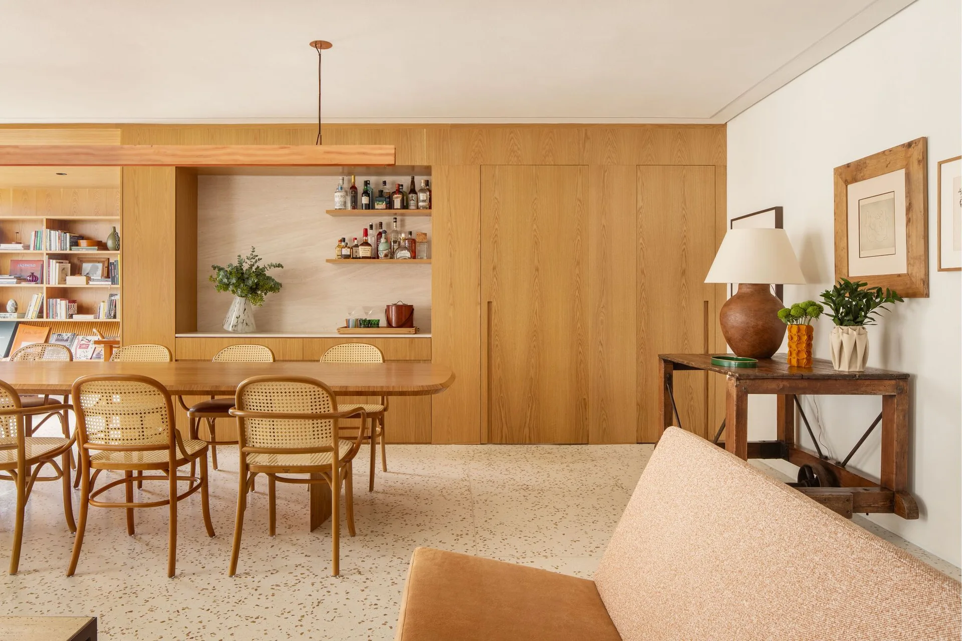A dining room features a wooden table, rattan chairs, and seamless wood wall cabinetry with a built-in bar over a speckled terrazzo floor.