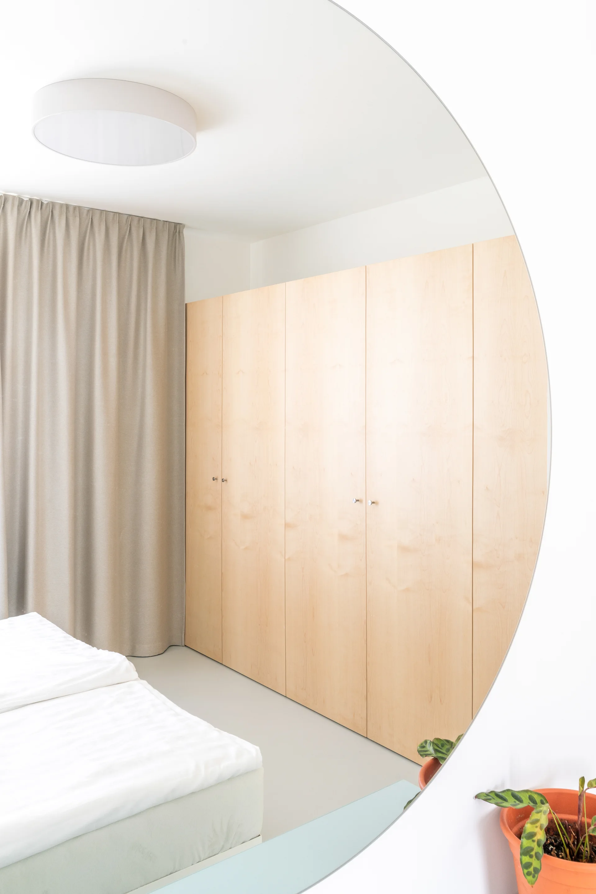A minimalist bedroom features a large light wood wardrobe, beige curtains, white bedding, and a reflection in a large circular mirror.